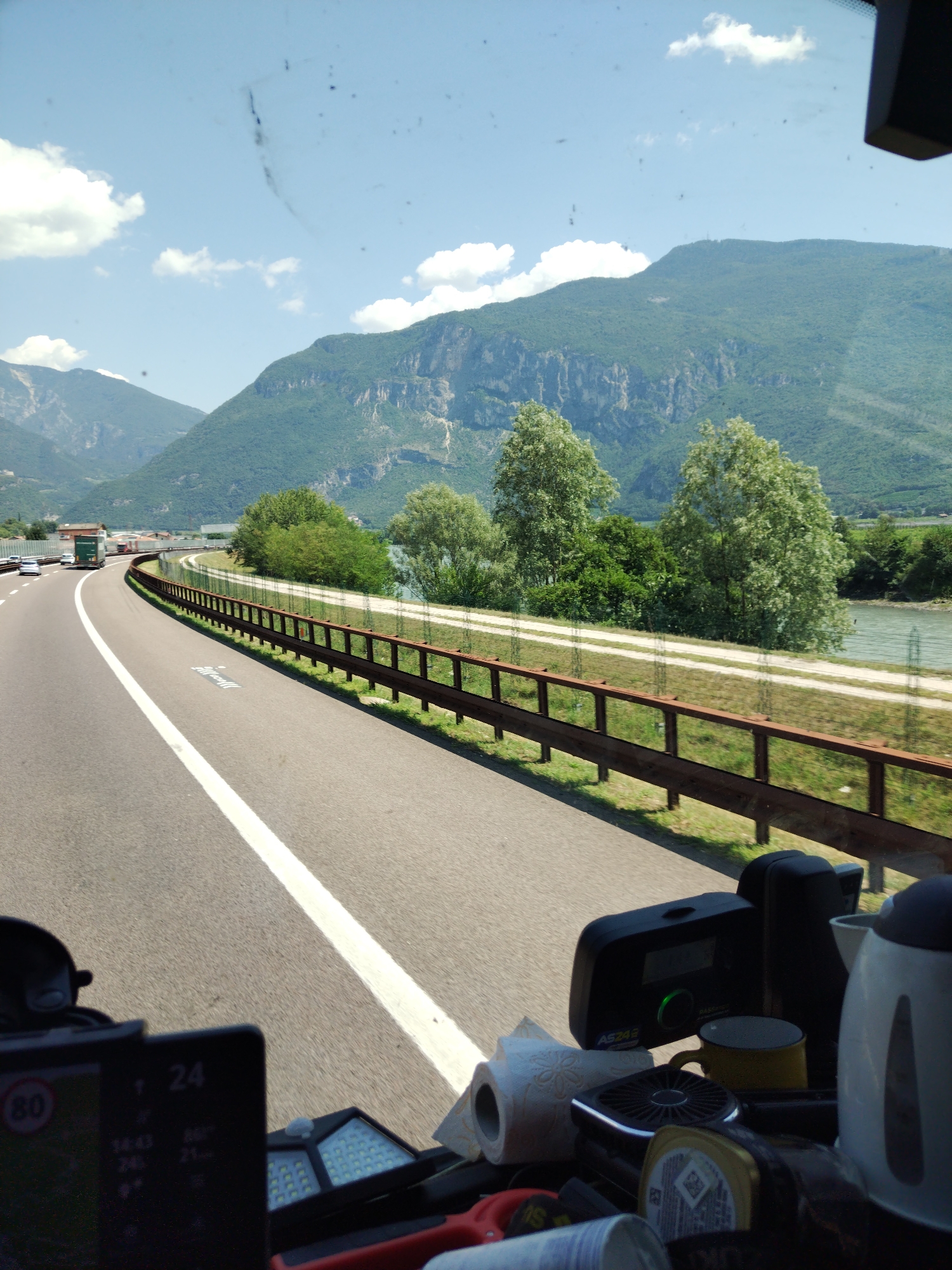 The image captures a view from inside a vehicle, likely a truck or RV, looking forward through the windshield. The vehicle is traveling on a multi-lane highway with a white line marking the lanes. The highway curves to the left, bordered by a metal guardrail and green vegetation. Beyond the guardrail, a river is visible, and further in the distance, lush green mountains with rocky outcrops rise against a clear blue sky with scattered white clouds. The mountains appear to be in the vicinity of Volano, Italy, as suggested by the terrain and foliage.
In the foreground, inside the vehicle, several objects are visible on the dashboard. A GPS device or navigation unit with a screen displaying "80" within a red circle (likely a speed limit indicator) and other digital information is prominent on the left. To the right, other items include a roll of toilet paper, a yellow mug, a black electronic device with a green light, and a white electric kettle. The dashboard also shows some smudges or dirt on the windshield.
The overall scene depicts a journey through a scenic mountainous region, likely on a sunny day, with the driver's perspective focused on the road ahead and the surrounding natural landscape.