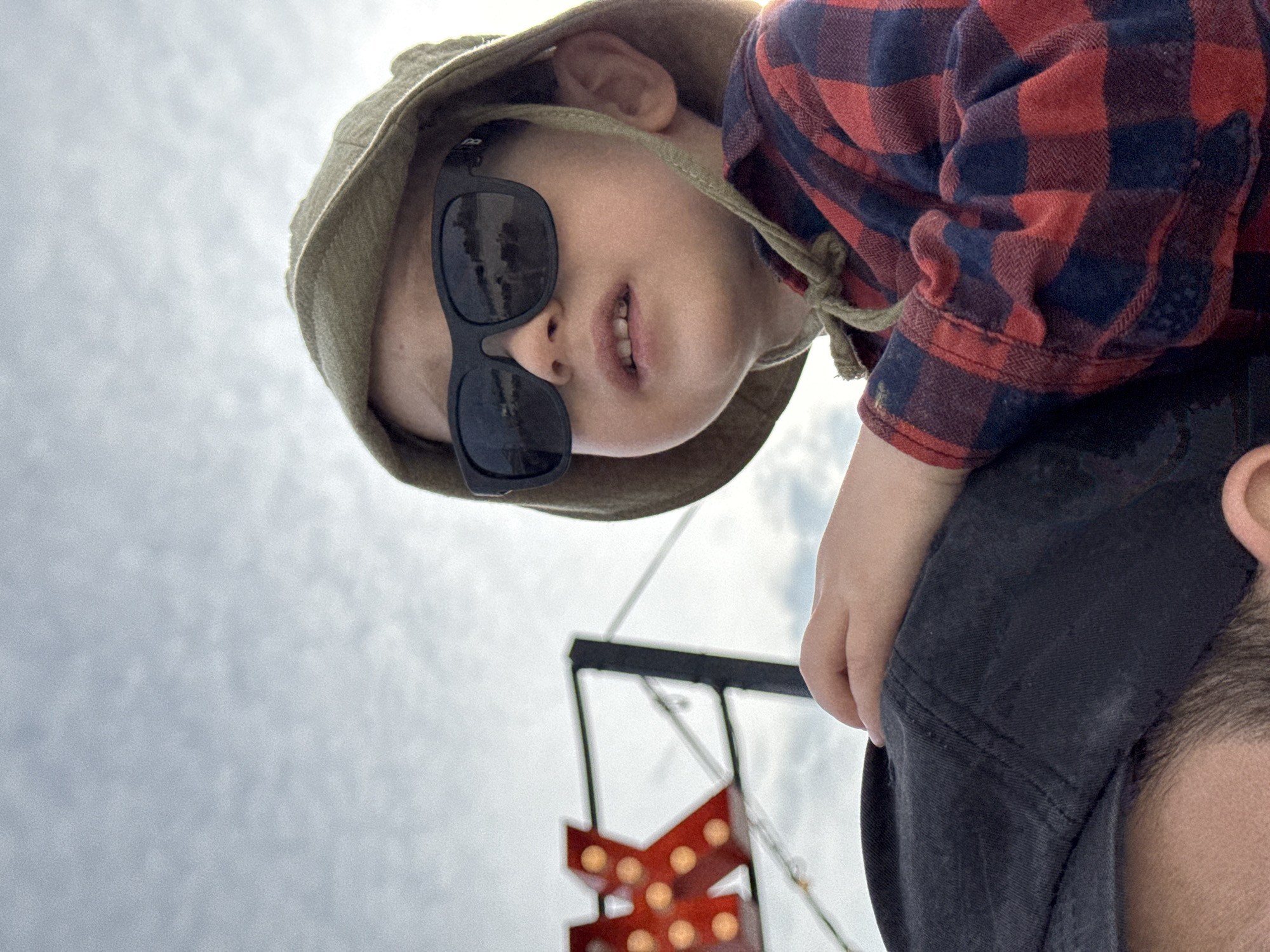 A low-angle, outdoor shot shows a young child wearing sunglasses and a bucket hat riding on someone's shoulders. The child is wearing a red and blue plaid shirt. The person carrying the child is wearing a dark-colored cap. In the background, partially obscured, is a structure with illuminated orange lights, potentially part of a sign. The sky is cloudy. The location is Sochi, Russia.