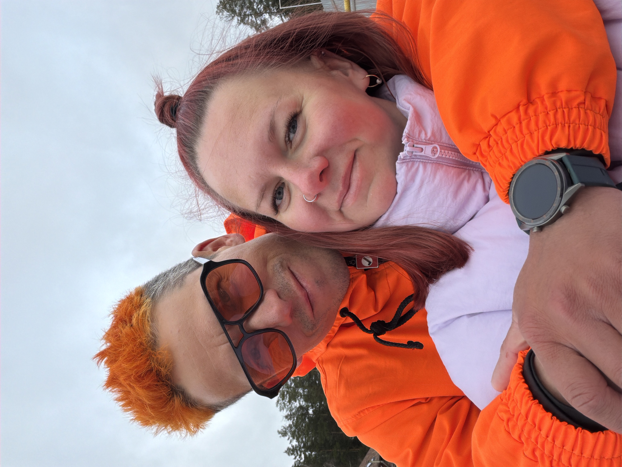 A close-up, angled portrait captures two individuals, a man and a woman, posing together outdoors in Chaikovsky, Russia, against an overcast sky. The woman, positioned slightly above and to the left, has reddish-brown hair styled in a messy bun on top of her head. She wears a silver hoop nose ring and earrings, and looks towards the camera with a subtle smile. She is dressed in a vibrant orange jacket over a lighter, possibly white or light pink, inner layer with a visible zipper.
The man is positioned below the woman, partially obscured. He has distinctly bright orange, dyed hair, with natural grey/white hair visible at his temples. He wears orange-tinted sunglasses with a prominent black frame and has a slight smirk on his face. He is also wearing a matching orange jacket with elasticated cuffs. A dark-faced smart watch with a grey band is visible on his left wrist. The background is dominated by a light, cloudy sky, with a few dark coniferous trees visible in the distance. The overall impression is one of a relaxed and affectionate moment between the two.