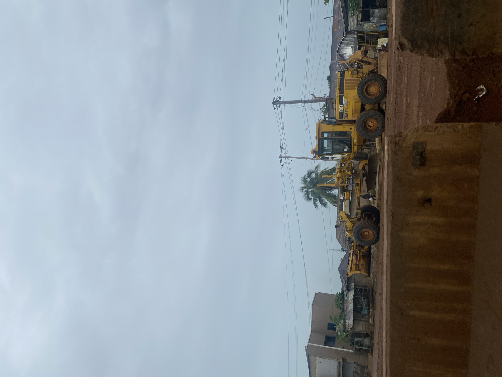 This image depicts a construction site in Keffi, Nigeria, under an overcast sky. A yellow Caterpillar 140H motor grader, a heavy construction vehicle, is the central focus, positioned on a dirt surface. The grader has large rear wheels and a blade in the front, suggesting recent or ongoing work. Behind the grader, several structures are visible, including a partially completed building with exposed framing, a small shack with a corrugated metal roof, and other buildings with similar roofing materials. The environment appears to be rural or semi-urban, with a palm tree visible to the left of the grader and utility poles and power lines stretching across the background. The foreground is dominated by a rough, reddish-brown concrete barrier. The scene suggests a developing area where infrastructure work is being undertaken. No individuals are clearly visible in the image, though a small figure in dark clothing can be seen walking to the right of the grader. The overall impression is one of ongoing construction and a somewhat unkempt environment.