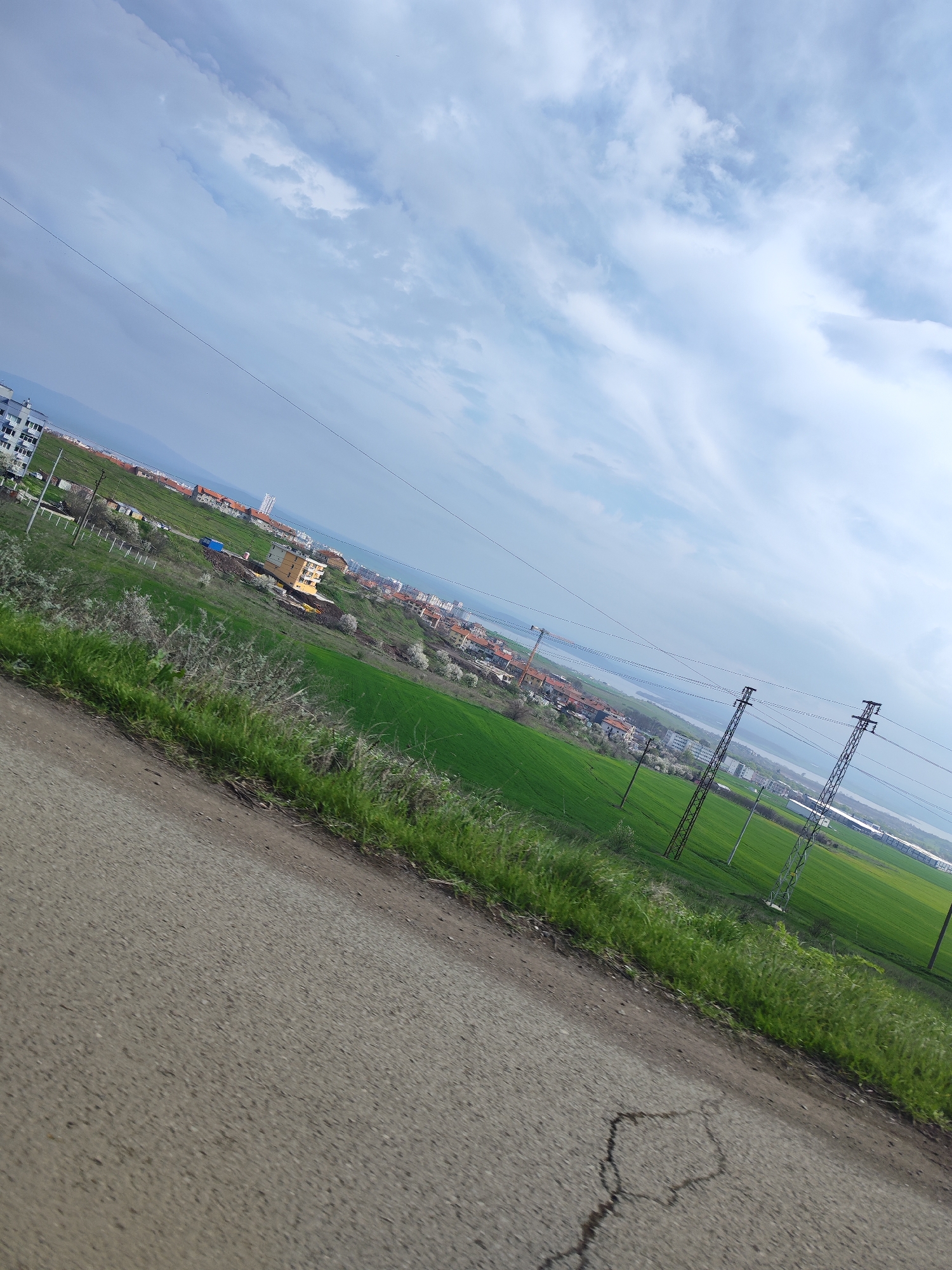 The image is taken at an oblique angle along an asphalt road, looking towards a settlement in the city of Burgas, Bulgaria. A field of green grass occupies the middle ground, featuring several electrical transmission towers. Beyond the field, a body of water is visible, bordered by buildings that comprise a distant skyline. The sky overhead is a mix of blue and white clouds. The focus is on the landscape and infrastructure rather than any specific human activity. The foreground is dominated by the road surface, marked by cracks and patches. The scene is indicative of a rural-urban fringe area.