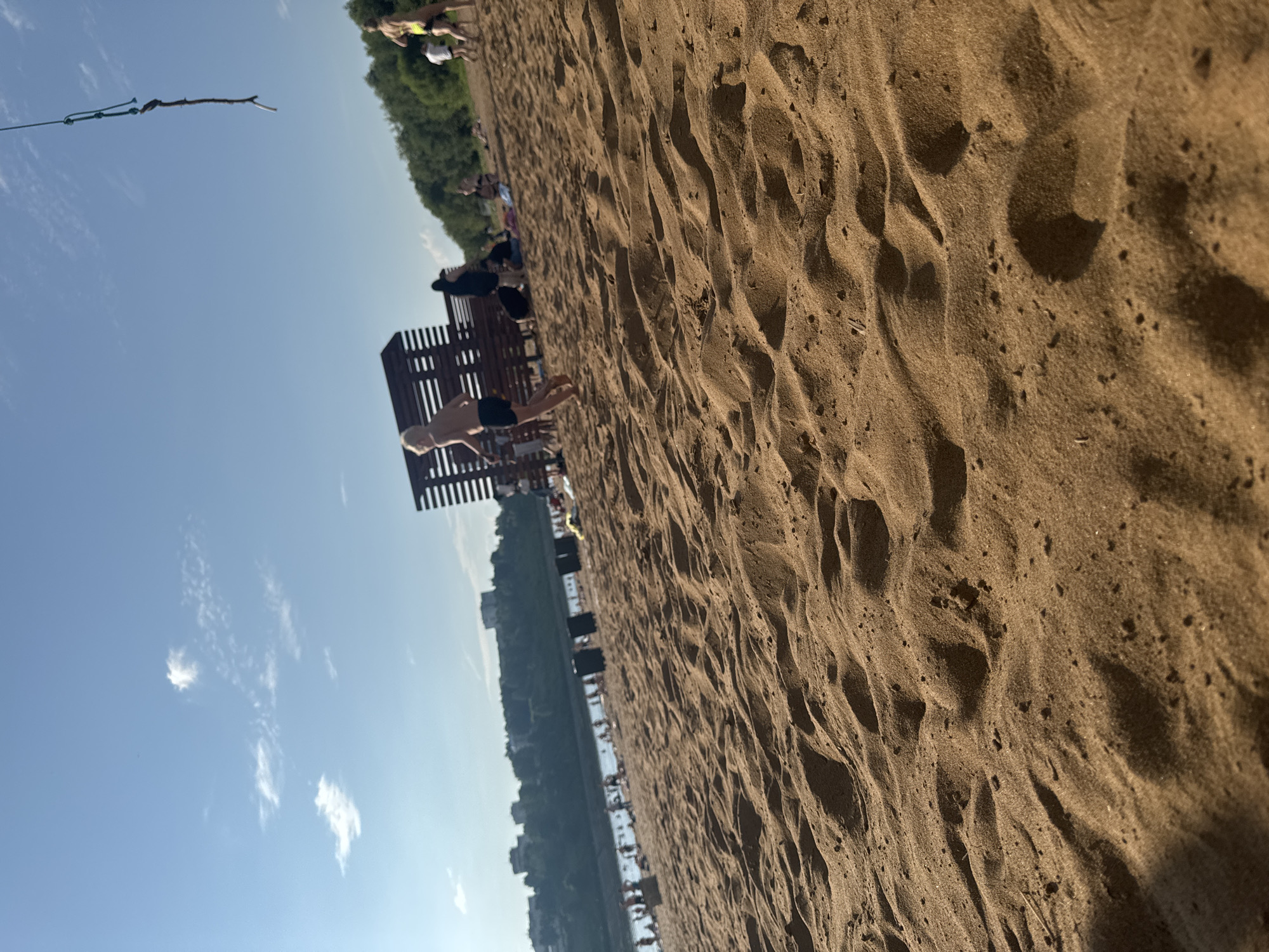 The image depicts a sandy beach on a sunny day. In the foreground, the sand is textured with ripples and shadows. In the middle ground, several people are present, some sitting or lying on the sand, while others are walking. A young boy in swim trunks is running towards the left of the frame. Behind the beachgoers, a wooden structure, possibly a changing room or a sunshade, stands. Further in the background, a body of water is visible, with more people near its edge. A tree-lined bank with buildings can be seen across the water, indicating an urban or semi-urban environment. The sky is blue with scattered white clouds. A piece of wood tied to a rope hangs from the top right corner of the image, suggesting a possible swing or a recreational setup. The scene appears to be a public beach, likely a popular spot for recreation in the city of Kirov, Russia, given the presence of a river or large body of water and urban development along its banks.