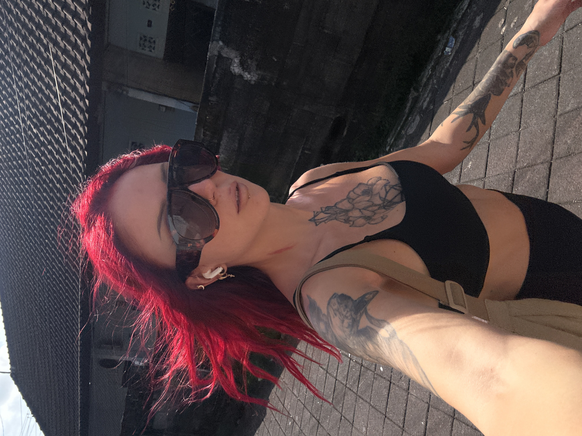 A woman with red hair and sunglasses is depicted in an outdoor selfie. She has tattoos on her arm and chest. She wears a black top and a beige bag is visible. The background shows a building and a tiled street. The scene is North Kuta, Indonesia.