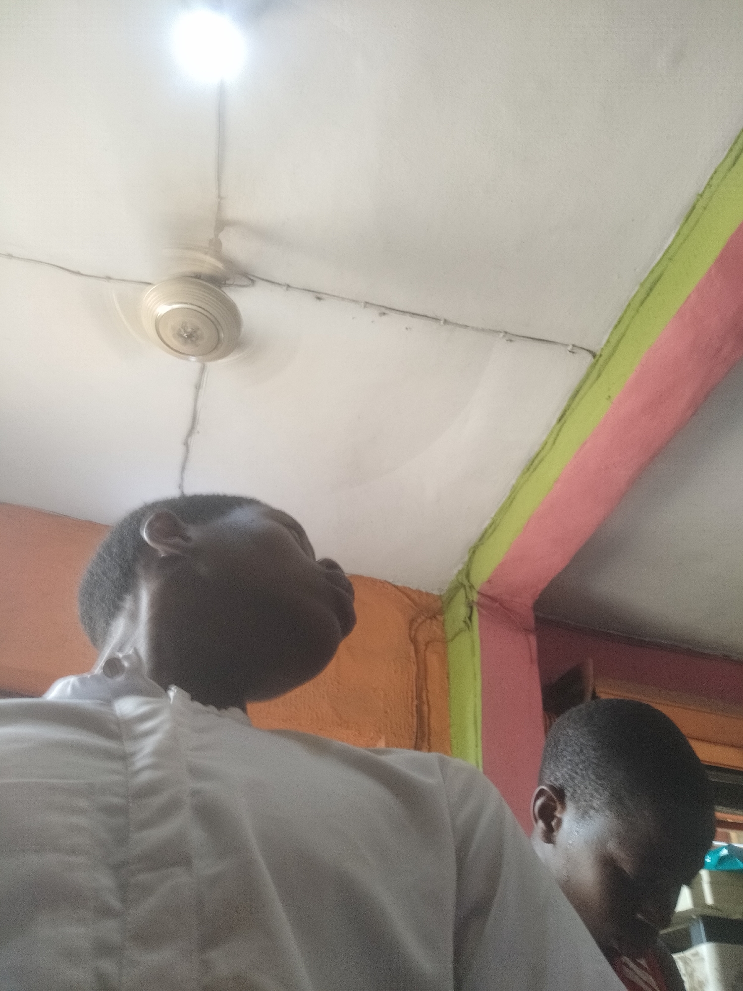 The image shows an indoor scene in Shangisha, Nigeria. Two dark-skinned individuals are visible, one prominently in the foreground wearing a white shirt, and another partially visible to the right. The background features a white ceiling with a rotating ceiling fan and a light fixture. Walls are painted in a combination of orange, green, and pink. The overall lighting is bright, possibly from the overhead light. The subjects appear to be indoors, possibly in a residential or commercial space.