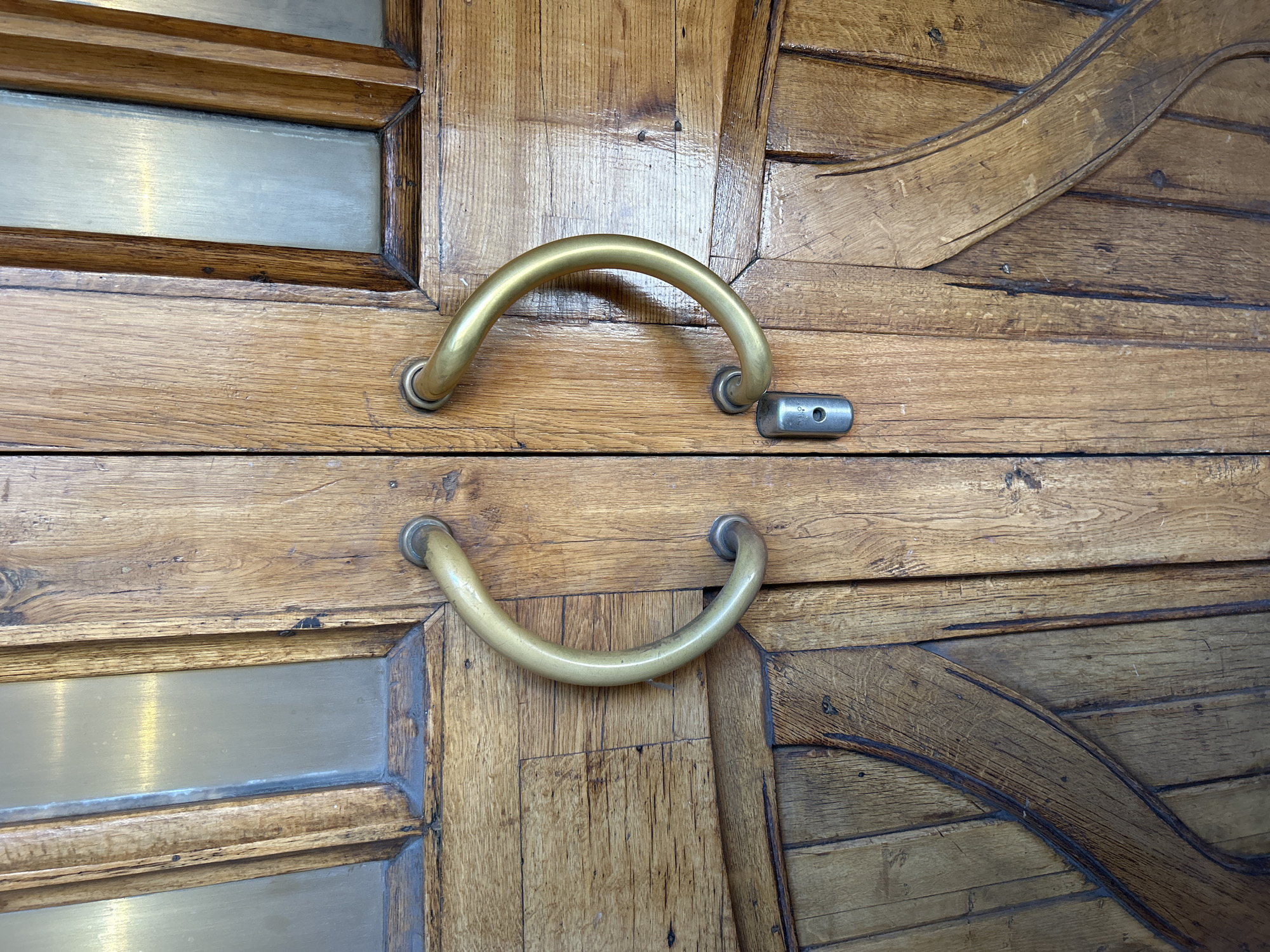 The image shows a close-up, eye-level view of a pair of ornate, wooden double doors. The doors are made of planks of light brown wood with visible grain and some natural imperfections. Each door features a rectangular pane of what appears to be frosted or opaque glass at the top, framed by the same wood. Centered on each door is a large, curved, brass door handle, resembling a "C" shape, attached with two circular mounts. Below the handles, in the center seam between the doors, is a small, rectangular, silver-colored lock mechanism with a single visible screw. The overall impression is one of vintage elegance. The setting is not specified in the image or metadata.