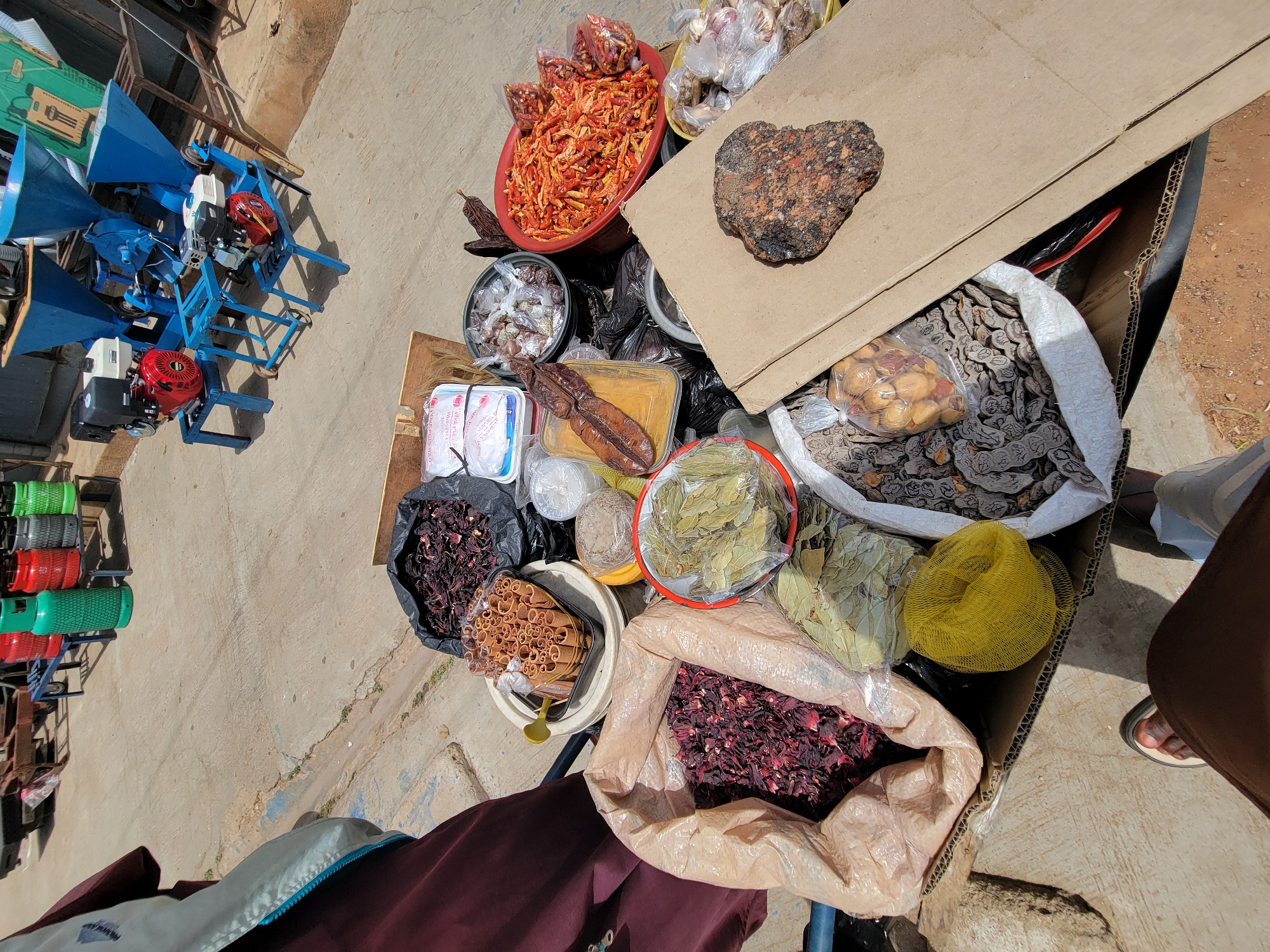 The image, presumably taken in the city of Jos, Nigeria, shows a collection of goods displayed for sale, likely at a local market. The goods are arranged in a disorganized manner, seemingly packed within a large cardboard box and various containers on the ground. The items include dried red peppers in a red bowl, spices, dried goods in plastic bags, and what appear to be medicinal herbs in various containers. There's a large, dark, heart-shaped object resting on a piece of cardboard. Also, there are three blue machines with red engines and a collection of colorful green and red plastic containers standing to the left. A person wearing brown pants and a sandal is visible near the box and a light-colored jacket is visible in the lower left. The background appears to be a concrete surface, possibly a street or sidewalk.