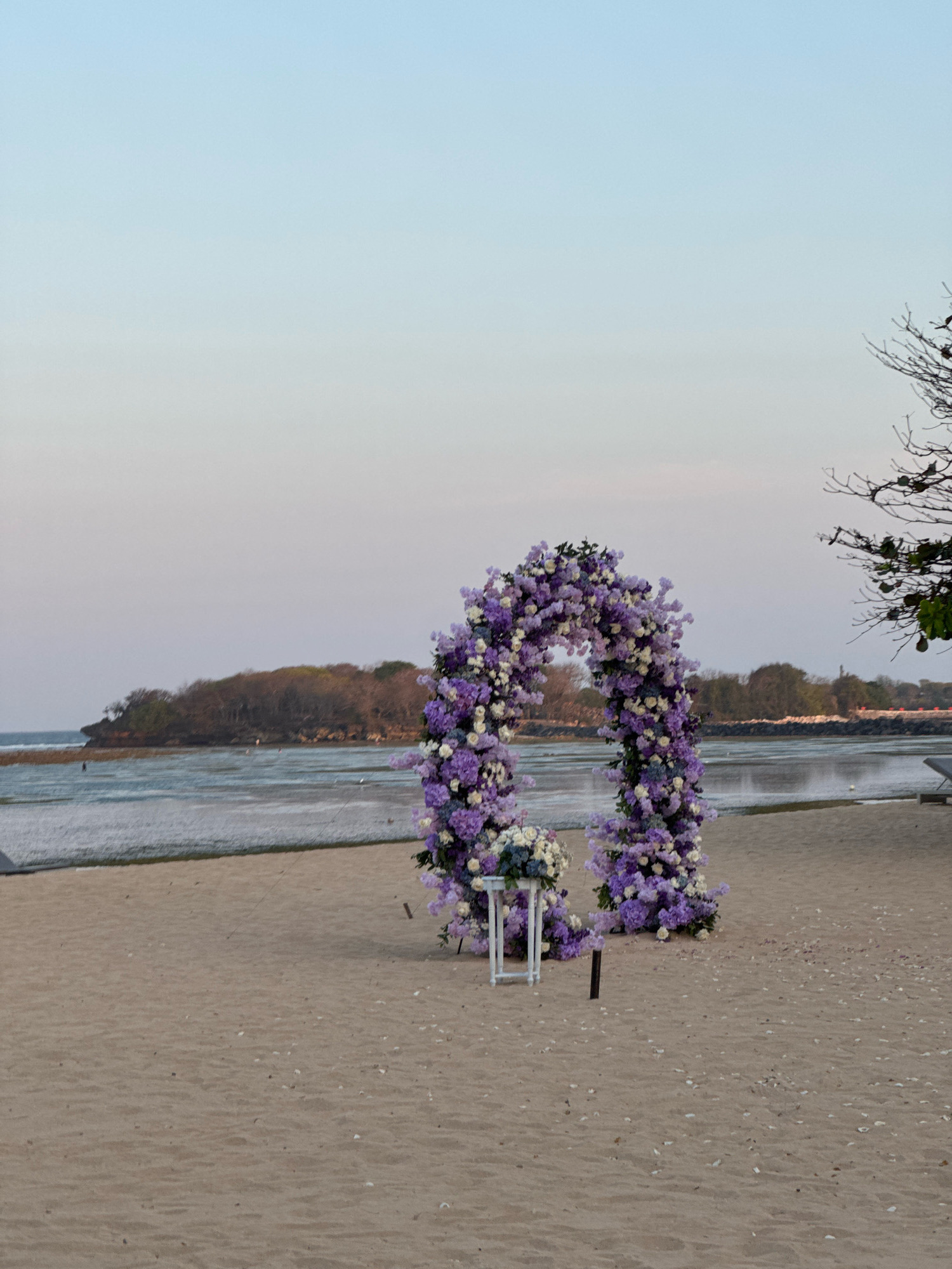 This is a medium shot, eye-level, outdoor photograph of a wedding arch decorated with purple and white flowers on a sandy beach. The arch is in the center of the frame, forming a semi-circle. A small white stand with a bouquet of flowers is placed in front of the left side of the arch. The beach is tan sand with some scattered white petals. The ocean is visible in the background, with a calm body of water, and a distant island with trees. The sky is a soft gradient of light blue and pale pink, suggesting it is either sunrise or sunset. A tree branch with minimal leaves is visible in the upper right corner of the frame. The location is a beach in Kuta Selatan, Indonesia. There are no people visible in the frame.