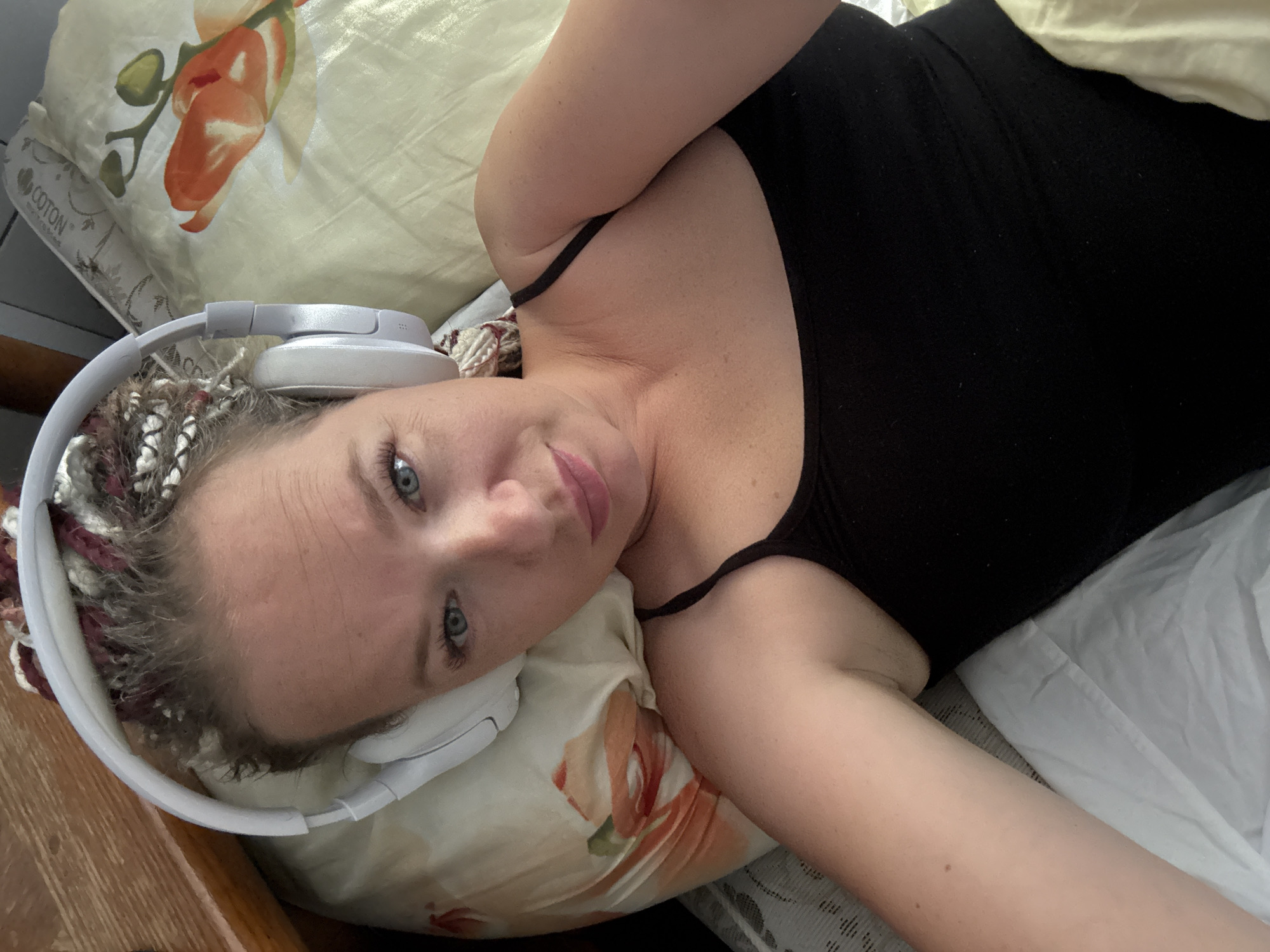 A woman with braided hair and blue eyes is lying on a bed, wearing white headphones and a black tank top. She is looking at the camera, with her head tilted slightly. The bed has a pillow with a floral pattern and a light-colored sheet. A wooden piece of furniture is visible to the left of the frame. The scene appears to be indoors, in a bedroom. There is no indication of the location being Sochi, Russia.
