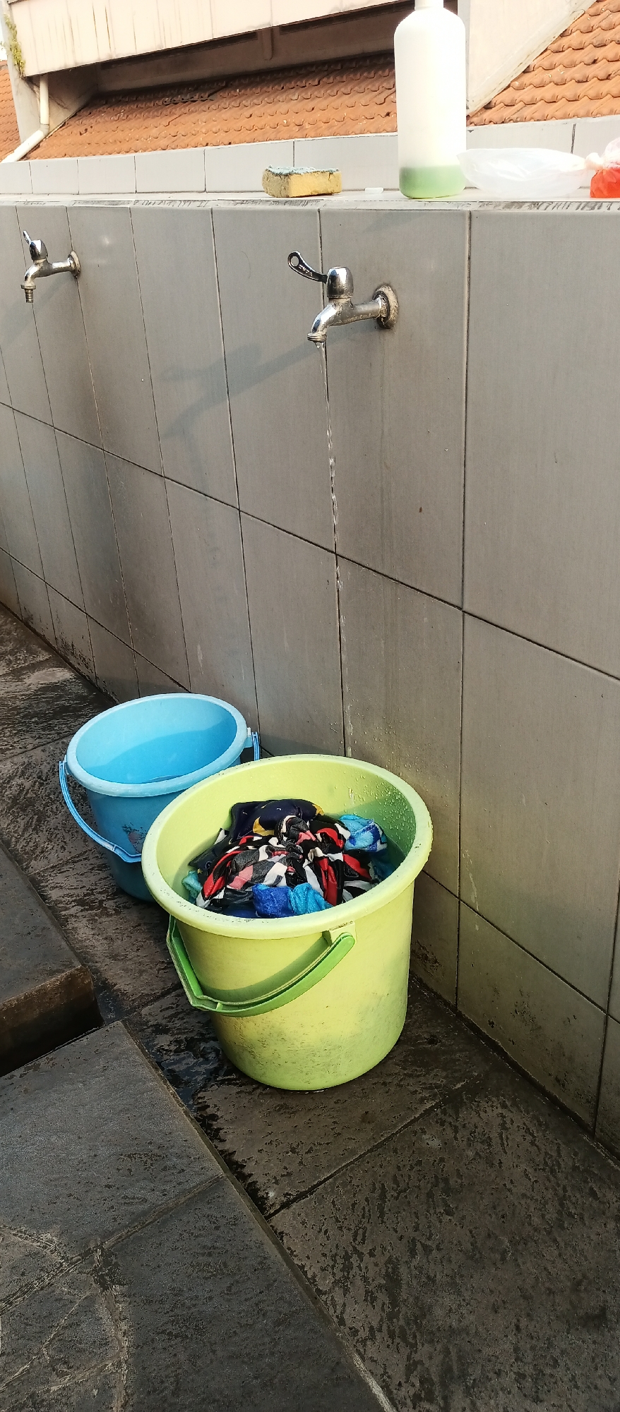 The image depicts an outdoor laundry area with two buckets on a tiled floor. One bucket, colored light blue, is empty and filled with clear water. Next to it, a larger, lime green bucket contains laundry submerged in water. A silver faucet on a tiled wall above the buckets is actively dispensing water into the green bucket. To the left, another similar faucet is visible, but it is not in use. On the tiled ledge above the faucets, a yellow sponge and a white bottle with green liquid are present. The background shows a portion of a building with a tiled roof. The location is identified as a laundry area in West Jakarta, Indonesia.