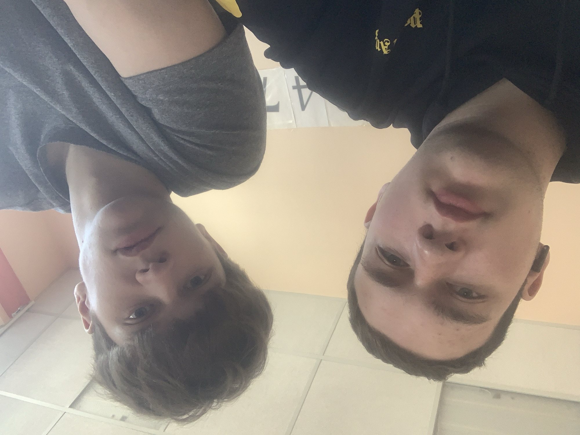 Two fair-skinned males are visible from a low-angle perspective, likely a selfie or similar self-shot. The male on the left is wearing a grey shirt and has brown hair. The male on the right is wearing a black hooded sweatshirt with a gold emblem and appears to have short brown hair. The background includes white tiles, a beige wall, and an area of red and orange coloration. A sign with the number "47" is also visible. Based on the context of the query (Severodvinsk, Russia), the location is likely an indoor space, potentially a school, gym, or locker room.