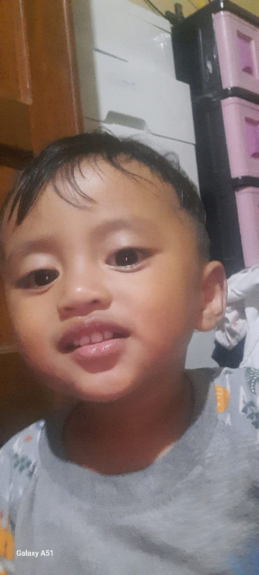 The image shows a close-up portrait of a young child, likely male, with dark hair and brown eyes. The child is wearing a light gray shirt with a printed pattern of palm trees and orange circular shapes. The background is out of focus, but appears to be an indoor setting. To the right of the child, there is a black plastic storage unit with pink drawers. To the left, a wooden surface is visible. The bottom left corner of the image contains the text "Galaxy A51", indicating the device used to take the photo. The overall impression is a candid, informal photograph taken in a domestic environment. The location is identified as Jatitengah, Indonesia.