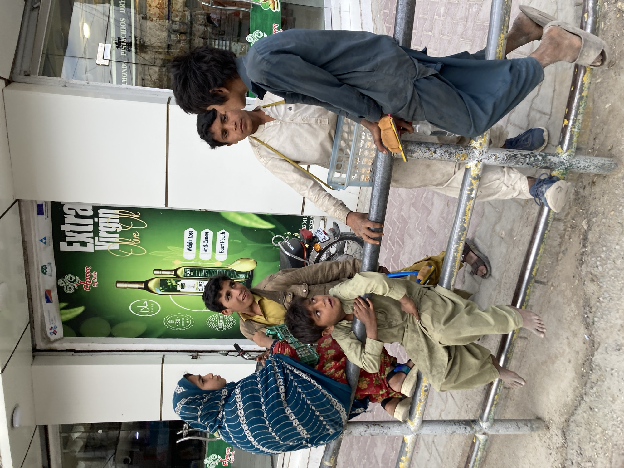 The image captures a scene in Quetta City Tehsil, Pakistan, featuring a group of individuals, including children and an adult woman in a blue patterned hijab, interacting with a metal structure that seems to be a form of street furniture, possibly used for sitting or resting.
On the left side of the image, there is a storefront with a sign promoting "Extra Virgin Olive Oil" from Chaman Dry Fruits. The sign highlights health benefits like weight loss, anti-cancer properties, and heart health. The products include olive oil bottles. The sign also includes a Halal symbol, ISO certification, and a guarantee label.
In the foreground, a woman sits while children are seen perched on the metal structure, some barefoot, with varying expressions and poses. One boy is dressed in a dark blue outfit and wears sandals. Another boy wears a beige outfit. Another boy wears a yellow shirt. A motorcycle is parked in front of the sign. There is a yellow sticker on the pipe which looks like it has a Pakistani mobile phone number.
The environment suggests an urban setting, with the store serving as a backdrop and the metal structure standing on a paved area, potentially a sidewalk.