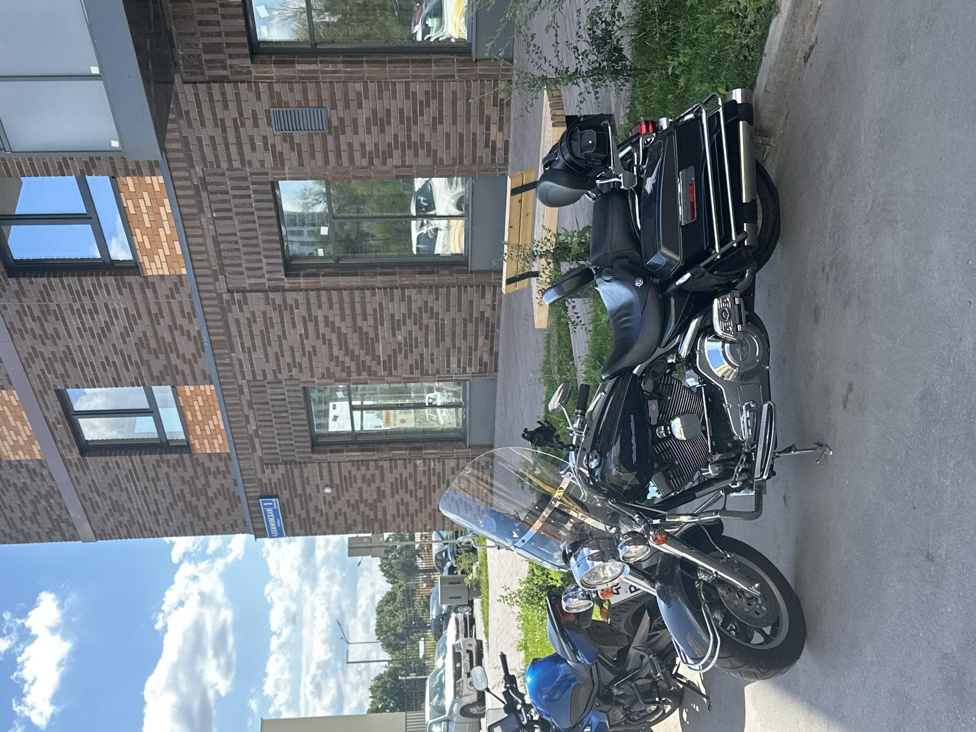 The image depicts a street scene in the city of Znamya Oktyabrya, Russia, on a sunny day with scattered clouds. In the foreground, two motorcycles are parked on a paved surface adjacent to a grassy area and a modern brick building.
The primary motorcycle in the foreground is a black cruiser with a large, clear windshield. It features a comfortable passenger seat with a backrest and a black bag attached to the rear rack. Chrome accents are visible on the engine, exhaust pipes, and other parts of the motorcycle. A blue motorcycle is partially visible to the left of the black one.
The building behind the motorcycles is constructed with dark brown and lighter orange bricks, featuring large windows and a modern aesthetic. A street sign with Cyrillic lettering, "ЗНАМЯ ОКТЯБРЯ 1" (Znamya Oktyabrya 1), is affixed to the building's facade. In the background, several cars are parked in front of the building. A wooden bench with metal legs is situated on a paved walkway in front of the building. The environment appears to be an urban or suburban residential area. There are no people visible in the image.