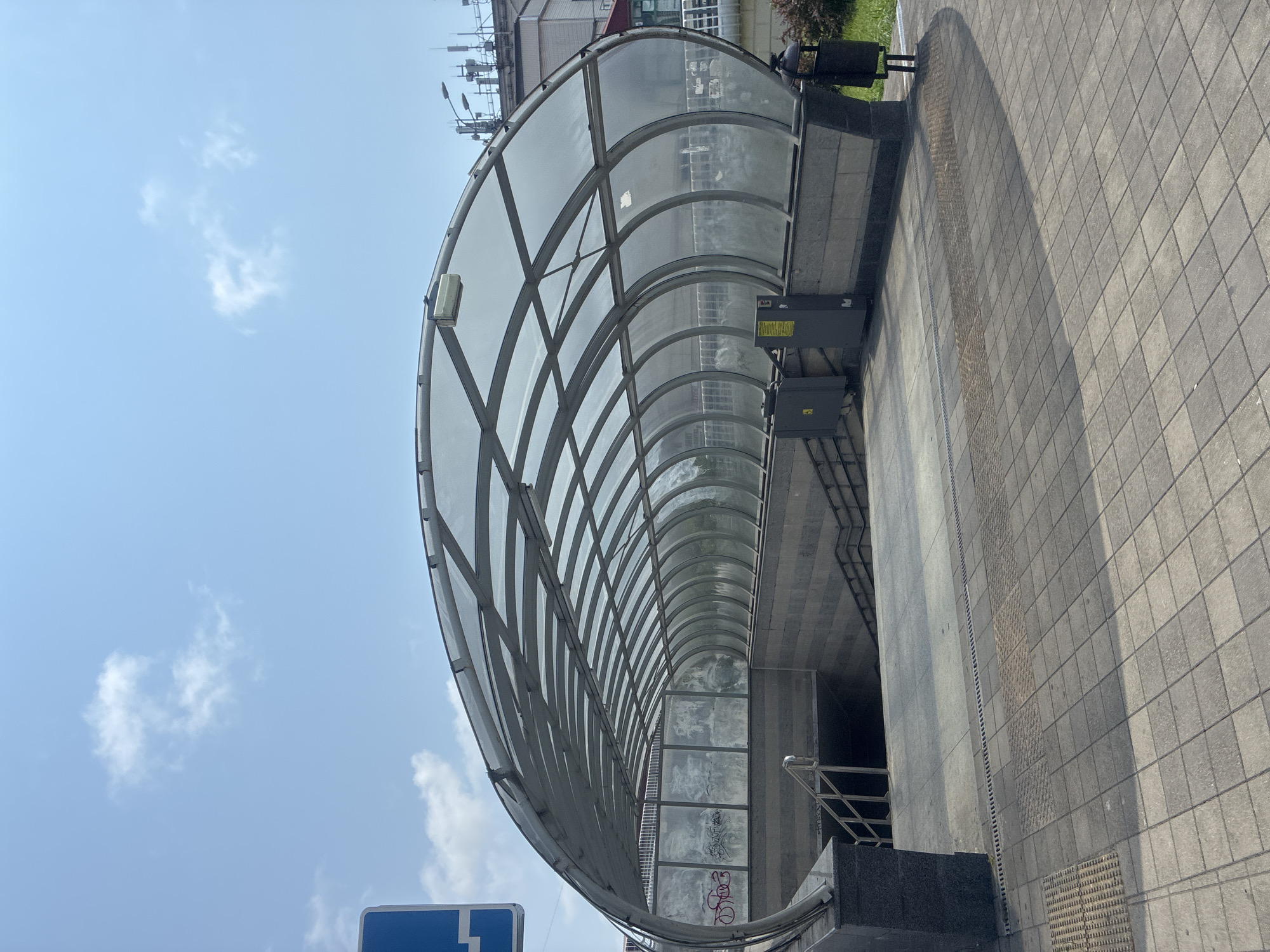 The image shows an overhead view of a modern subway entrance in Sochi, Russia. The entrance is covered by a translucent, arched canopy supported by a metal frame. The canopy is made of multiple panels that curve to form a tunnel-like passage. Stairs lead down into the subway station, flanked by stone walls and metal railings. The surrounding area is paved with square tiles, and a few pieces of street furniture, including a trash can, are visible. The sky above is blue with a few scattered clouds. The overall scene is clean and well-maintained, suggesting a functional and accessible public space.