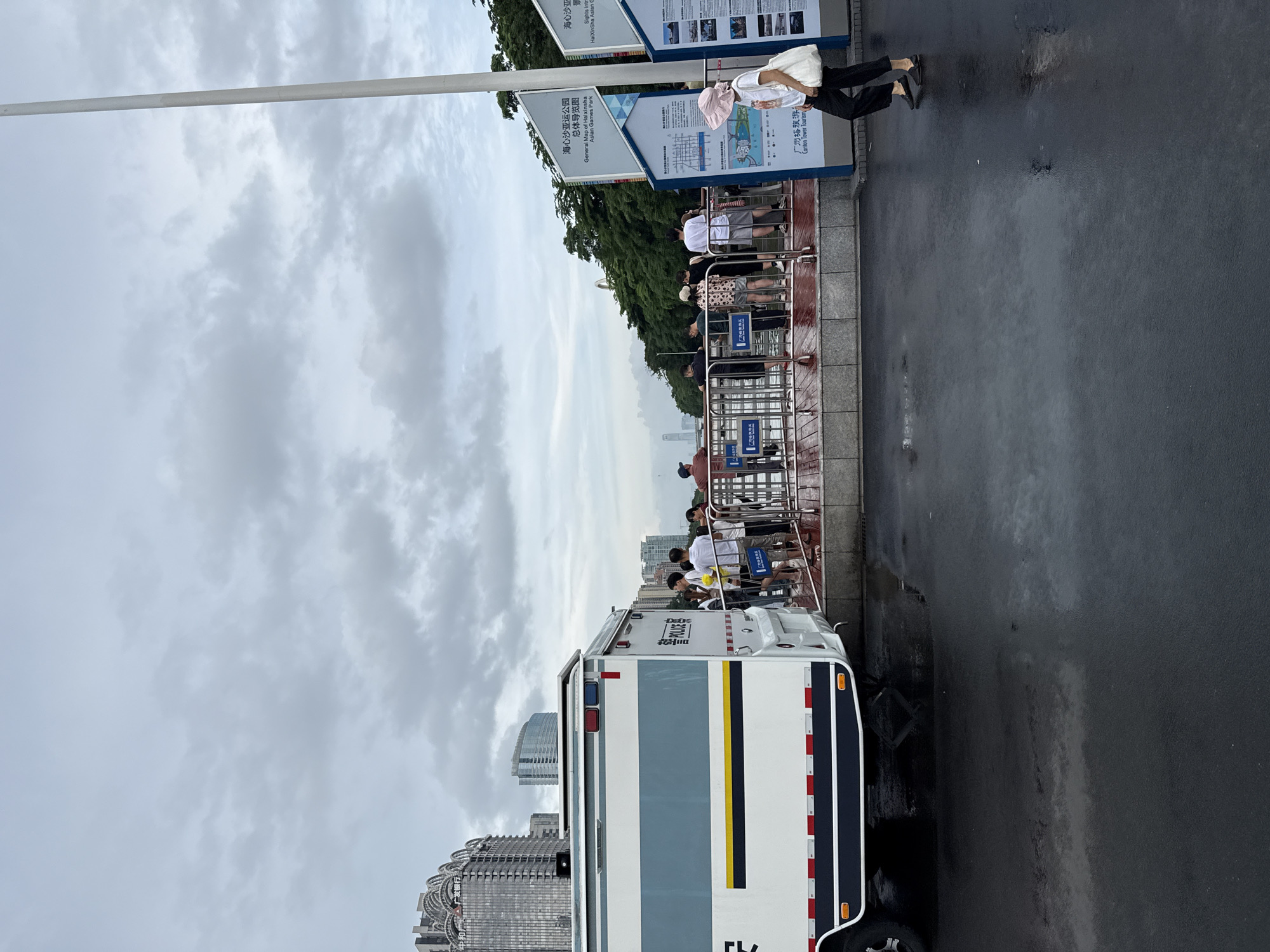 In Tianhe District, China, a white police vehicle with blue and gray accents is parked on a wet, asphalt surface near a metal railing. A group of people, appearing to be tourists, are gathered behind the railing, looking out towards a body of water. The sky is overcast. To the right of the railing, a woman wearing a pink hat and dark pants walks away from the camera. Behind the railing and to the right, informational signs are posted. In the background, tall buildings and lush green trees are visible.