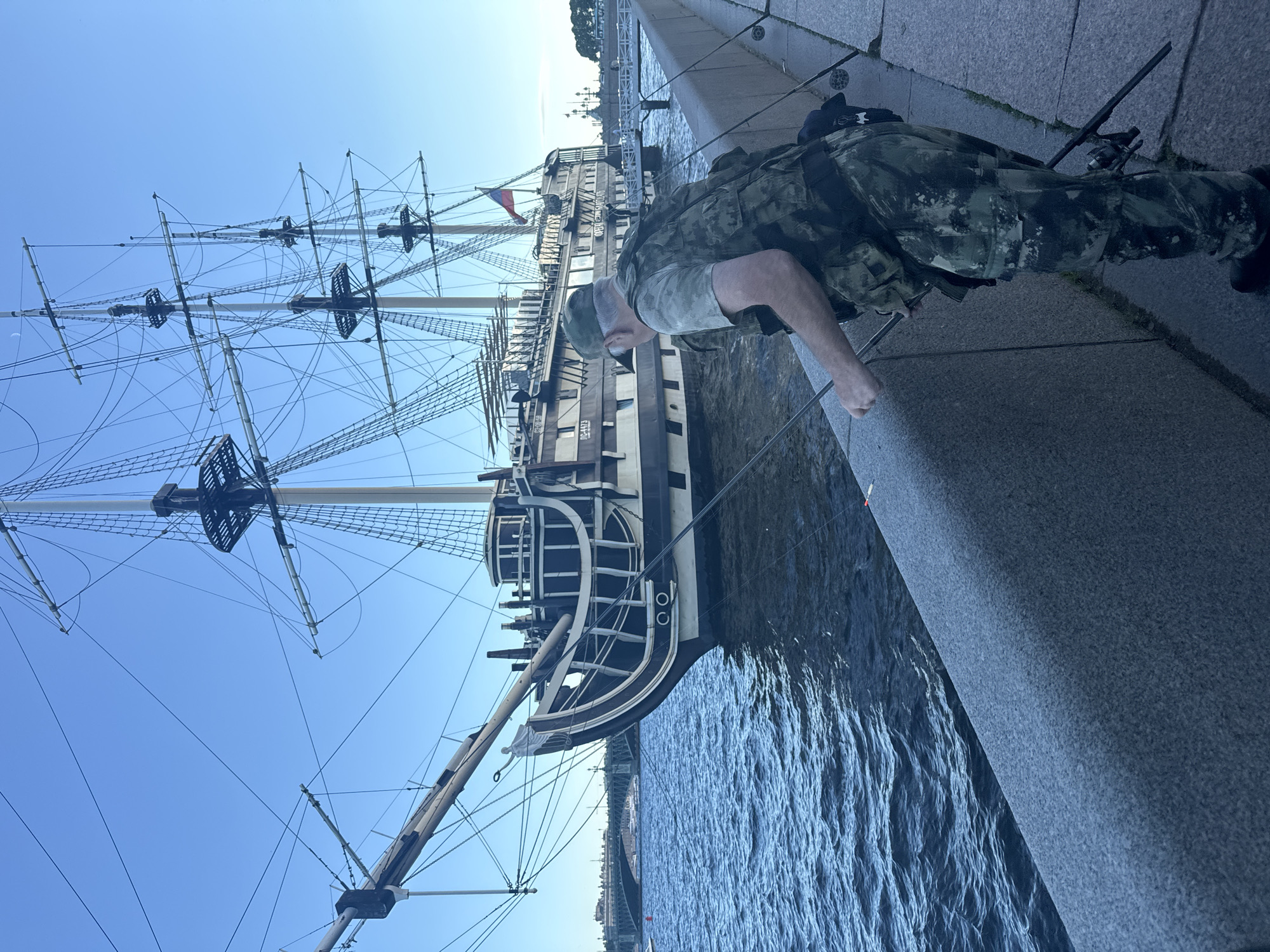 The image depicts a man fishing from a stone embankment in front of a large, historical-style sailing ship docked on a river. The ship, with its multiple masts and rigging, appears to be a replica or a preserved vessel. A Russian flag is visible on the ship's stern. In the background, a bridge and city skyline are discernible, suggesting an urban setting. The man is wearing camouflage fishing attire and is holding a fishing rod. The river is calm with gentle ripples on the surface. The sky is clear and blue, indicating fair weather. This scene is likely located on the Neva River in Saint Petersburg, Russia, given the architecture of the ship and the prominent bridge in the background, which resembles the Trinity Bridge.