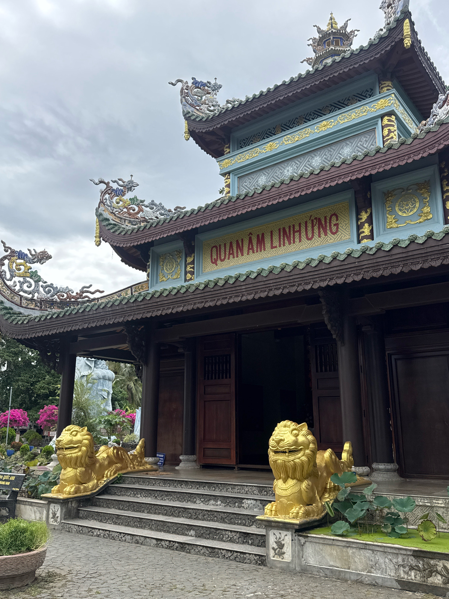 The image shows the entrance to a temple, likely a Buddhist temple, in Tuy An Nam, Vietnam. The temple features traditional Vietnamese architecture with ornate, multi-tiered roofs adorned with colorful dragon sculptures. A large sign above the entrance reads "QUAN ÂM LINH ỨNG" in gold lettering on a red background. Flanking the entrance stairs are two golden lion statues, a common motif in East Asian temples, symbolizing protection. The stairs themselves are made of stone, and in front of them, water features with lily pads are visible. The overall atmosphere is one of reverence and cultural significance, typical of such religious sites.