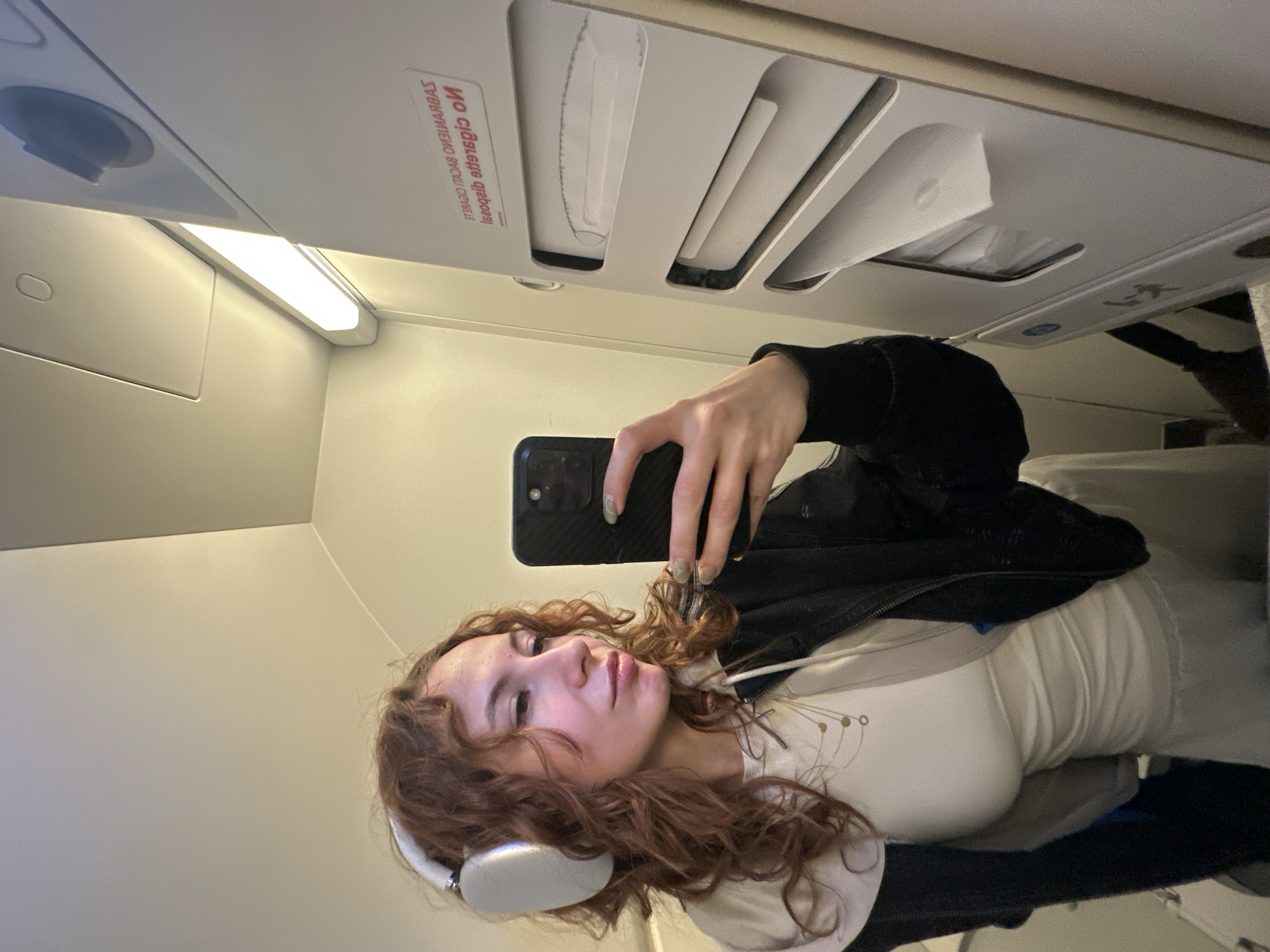 A young woman with curly, auburn hair is taking a selfie inside an airplane cabin. She is wearing white headphones, a white top, and a black jacket. She is holding a black smartphone in her right hand. The interior of the plane is visible, including overhead compartments with a "No Smoking" sign and a reading light. The location is not Podgorica, Montenegro; it is an airplane.