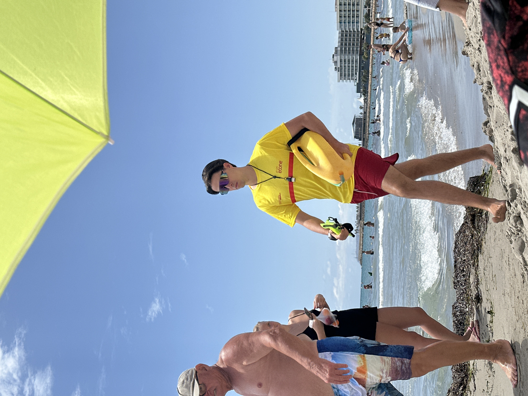 A medium shot shows a man in a yellow t-shirt and red shorts, wearing sunglasses, and holding a yellow rescue buoy on a beach. He is walking towards the right of the frame. In the background, other people are visible on the beach and in the water, along with buildings and a clear blue sky with some clouds. The sun is shining brightly, casting shadows on the sand. A portion of a yellow umbrella is visible in the top right corner of the frame.