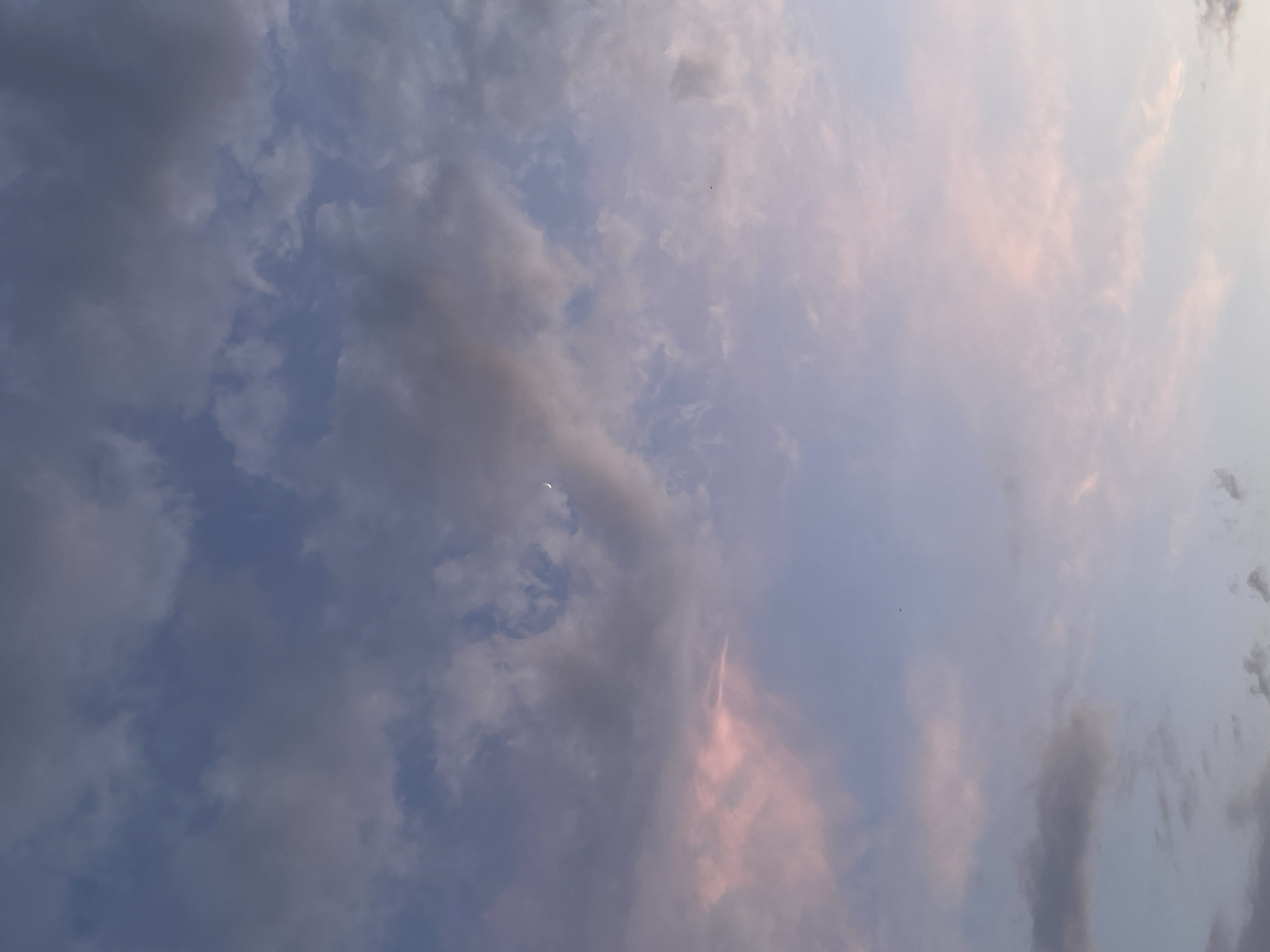 The image depicts a daytime sky with scattered clouds. The sky is a mix of blue and grey, with some clouds tinged with pink hues. A small crescent moon is visible in the left-center of the image. The photograph's perspective looks upwards. Location cannot be determined from the image, other than it is outdoors.