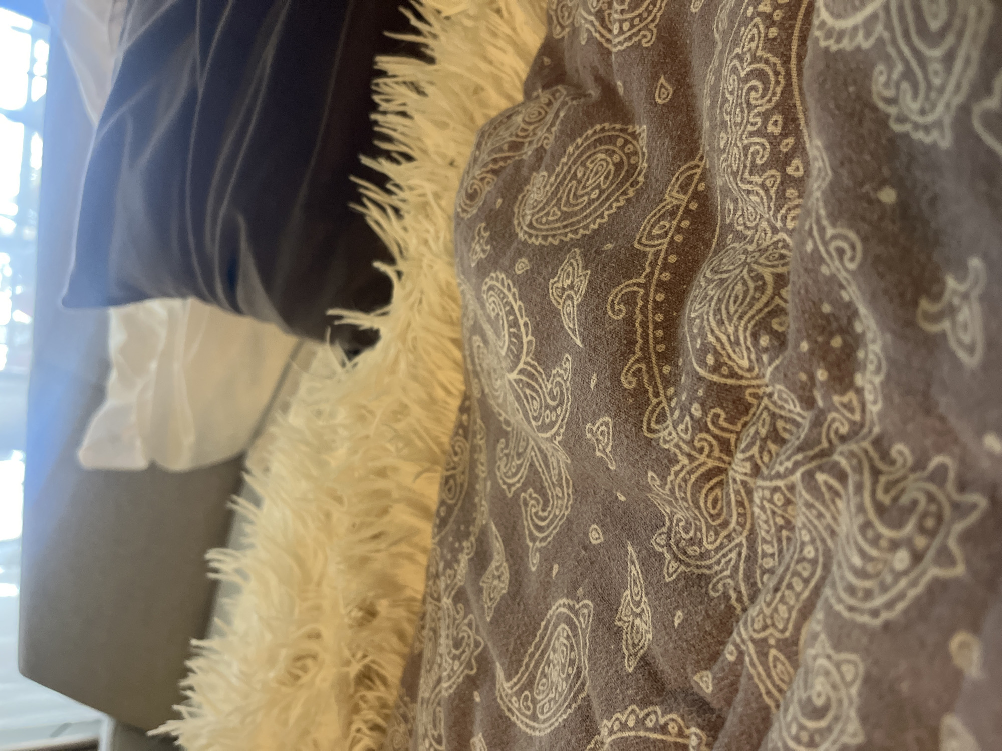 The image is a close-up shot of bedding with a paisley pattern in muted tones of brown and off-white. The fabric is soft and appears to be a blanket or comforter, with visible folds and wrinkles that create texture. To the left of the patterned fabric is a fluffy, cream-colored throw or faux fur blanket, also with visible texture and strand detail. Behind these blankets, a dark navy pillow is visible, its surface creased and soft. Further back, glimpses of white fabric and a gray upholstered surface suggest a domestic setting, possibly a sofa or bed. The lighting in the image is soft, with a gentle glow from what appears to be a window on the left side, indicating it might be daytime. The overall impression is one of comfort and relaxation. There is no visible text in the image.