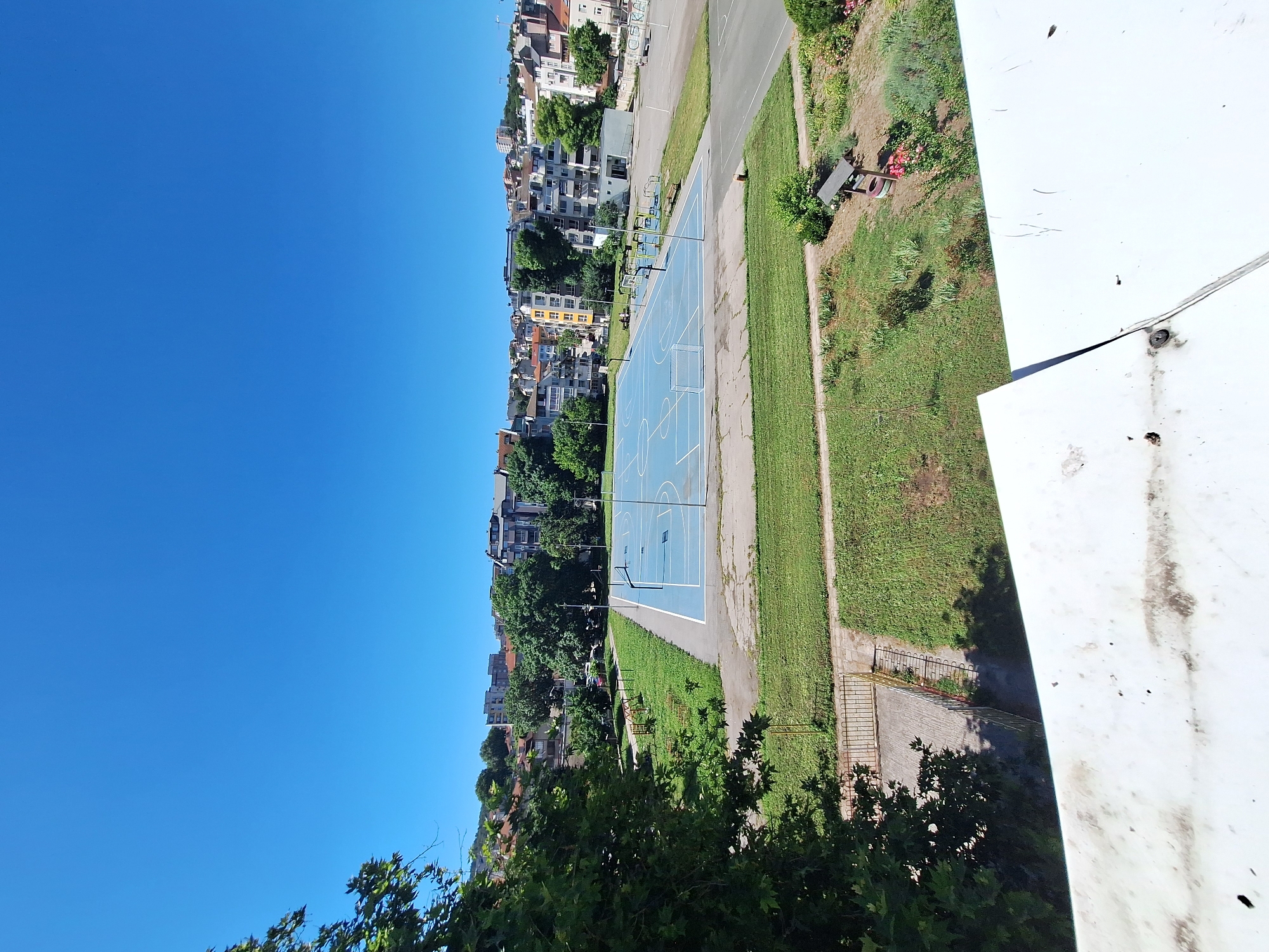 This is an exterior, high-angle, eye-level shot of a public recreational area on a clear, sunny day. The scene is set in the city of Varna, Bulgaria. Dominating the mid-ground is a bright blue, marked basketball court with white lines and basketball hoops. Adjacent to the court is a children's playground area featuring swings, slides, and climbing structures. The ground surrounding the court and playground is a mix of green grass, paved pathways, and concrete. In the background, the urban landscape of Varna unfolds, with a collection of multi-story residential buildings and mature trees. The foreground consists of the edge of a white, weathered roof. There are no people visible in the image.