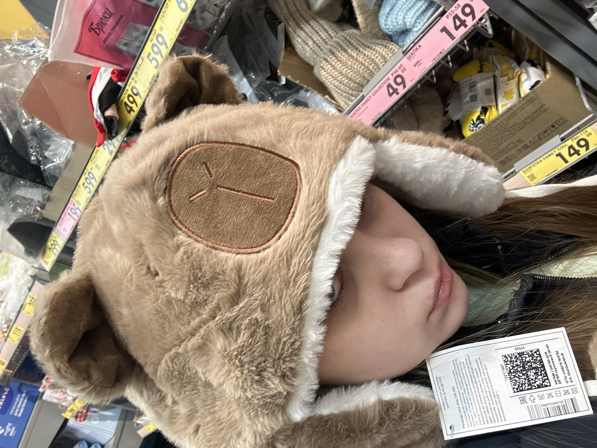 A person wears a brown bear hat in a store. Price tags indicate costs in Russian rubles: 49, 149, 499, and 599. A product label says "FAMILY COLLECTION," "MADE IN CHINA," and lists a Moscow address for AO "ARENA."