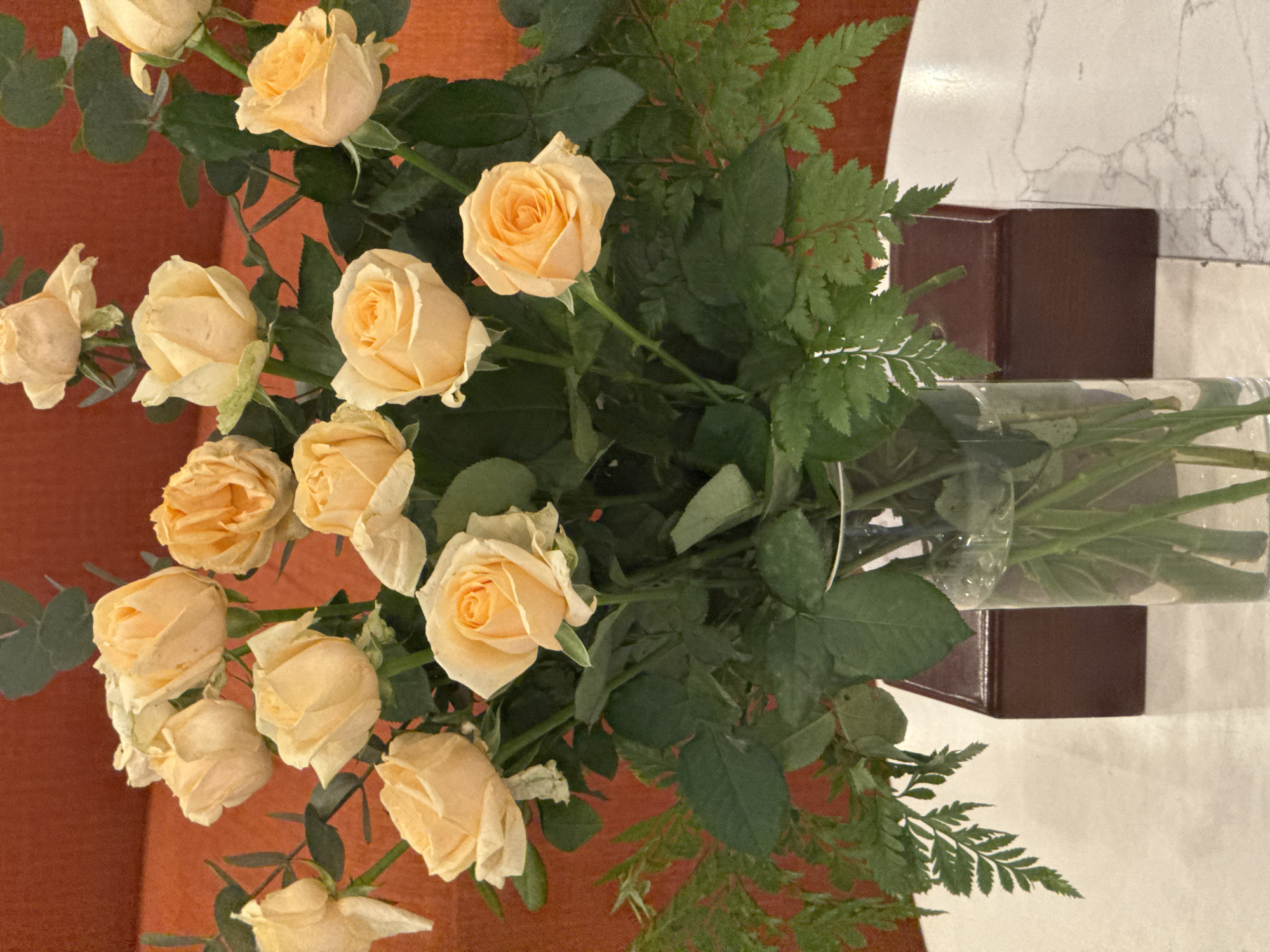 A bouquet of pale yellow roses with green leaves and ferns is displayed in a clear glass vase on a white marble-patterned surface. Behind the vase is a dark reddish-brown rectangular box, and further back is a textured orange wall. The scene does not provide any specific details that would indicate its location in Hanoi, Vietnam.