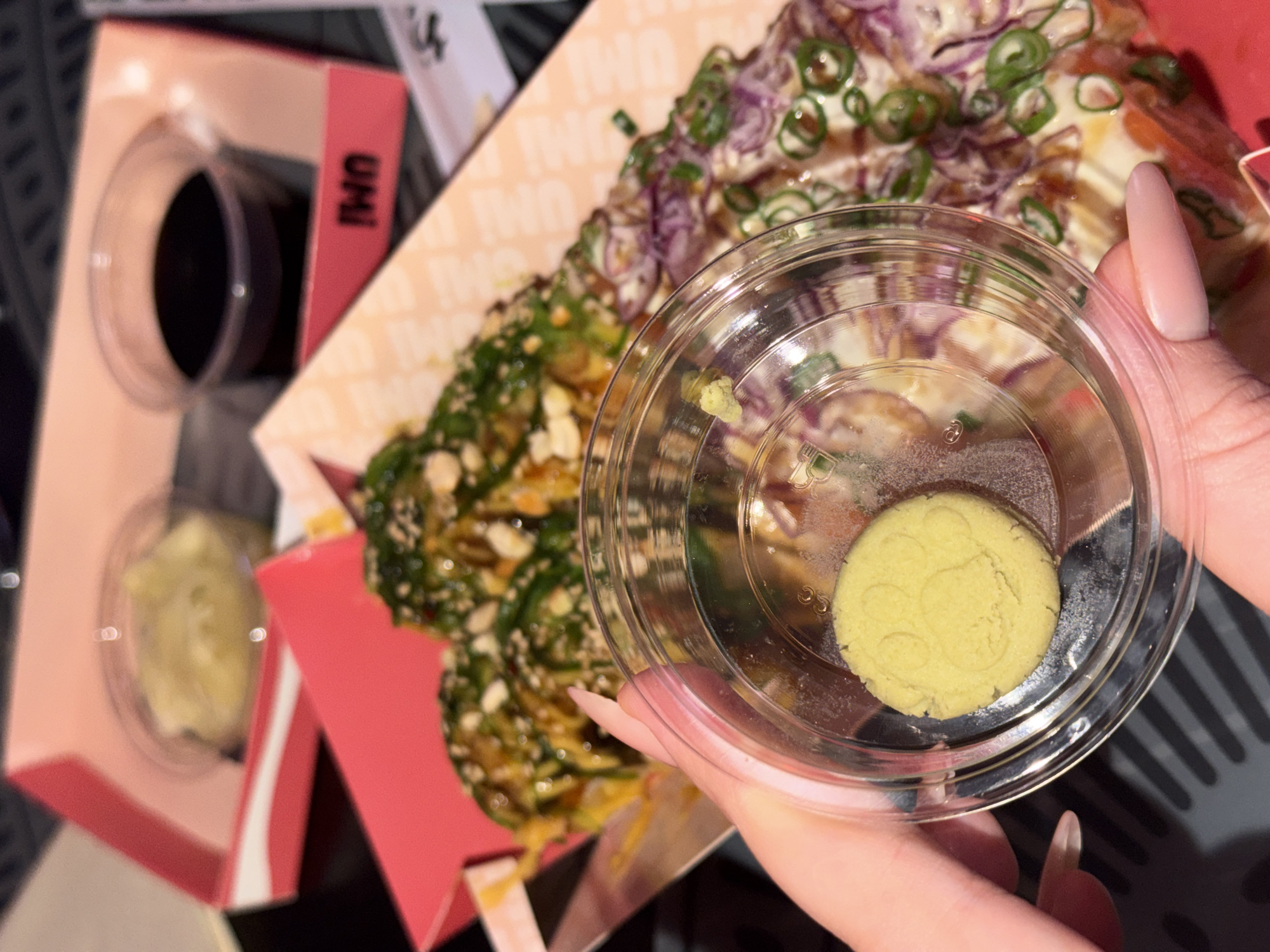 The image shows a close-up of a hand holding a small, clear plastic container with a round dollop of pale green wasabi paste inside. Behind the hand, out of focus, are parts of a takeout meal. This includes a portion of a dish with toppings like chopped green onions and shredded purple cabbage, and a container of soy sauce. The packaging suggests this meal is from "Umi," a restaurant. The takeout containers are pink and white. The overall scene suggests a casual dining or takeout setting. There is no indication that the photo was taken in the city of Belgrade, Serbia.