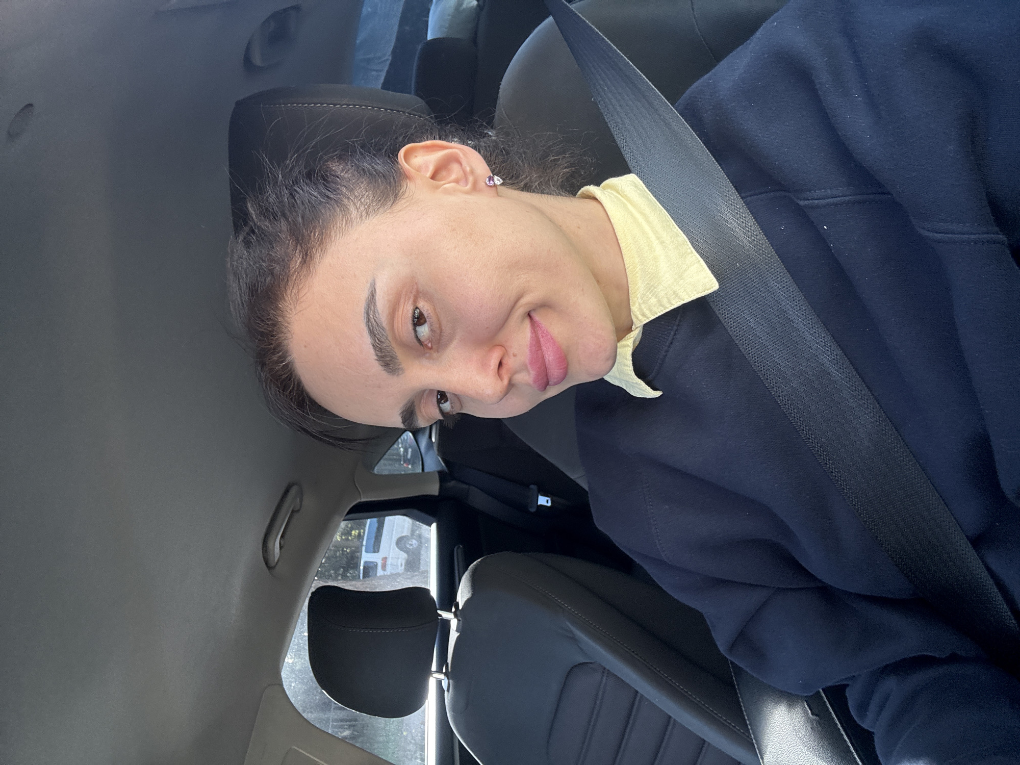 A woman is seated in the driver's seat of a vehicle. She is wearing a seatbelt, a navy blue sweater over a yellow collared shirt. Her hair is pulled back. The car's interior is primarily dark grey. Outside the vehicle's window, greenery is visible. The specific location in Tbilisi, Georgia, cannot be determined from the image.