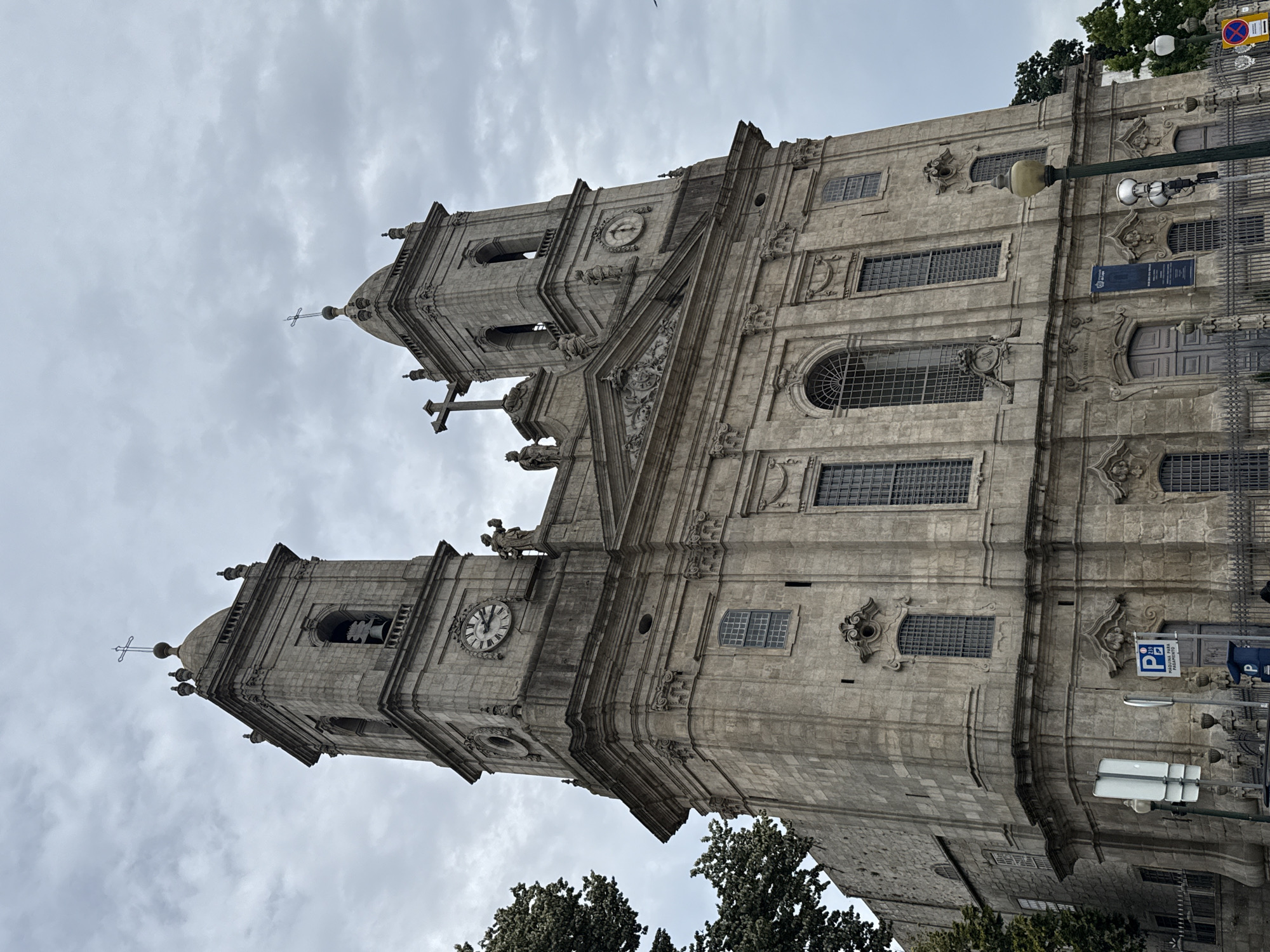 This is a vertical, low-angle, eye-level shot of the baroque facade of the Clérigos Church in Porto, Portugal. The church is constructed of light-colored stone and features two prominent towers with bell openings, a dome, and intricate architectural details like sculpted figures, ornate window frames, and decorative stonework. A large clock face is visible on the left tower. The sky is overcast with grey clouds, suggesting a cool or possibly rainy day. Trees are visible at the bottom of the frame, indicating an outdoor setting. A lamppost stands in front of the church, and a metal fence runs along the right side, suggesting a public space. A street sign with a parking symbol and text, as well as a blue sign with text, are visible near the fence. The overall impression is of a grand and historical religious building under a cloudy sky.