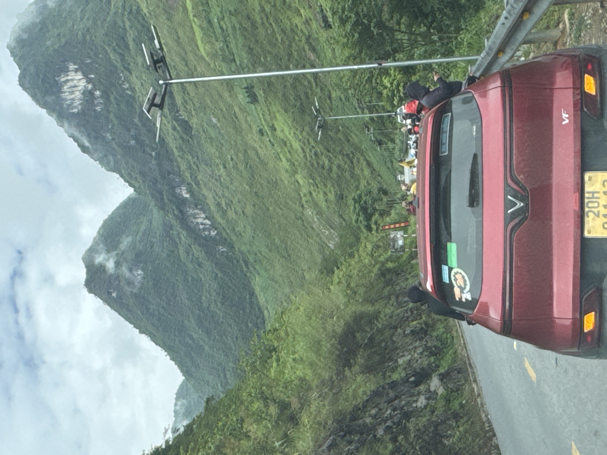 The image shows a red VinFast VF e34 car with the license plate 20H-041.13 parked on the side of a road, likely taken from another vehicle. The background features steep, green, cloud-covered mountains. The scene appears to be in a mountainous region, and according to the license plate number and general scenery, the location is most likely in the Yên Minh Commune, Vietnam. A crowd of people can be seen standing further up the road beyond the car. There is a guardrail along the side of the road, and streetlights extending into the scene.