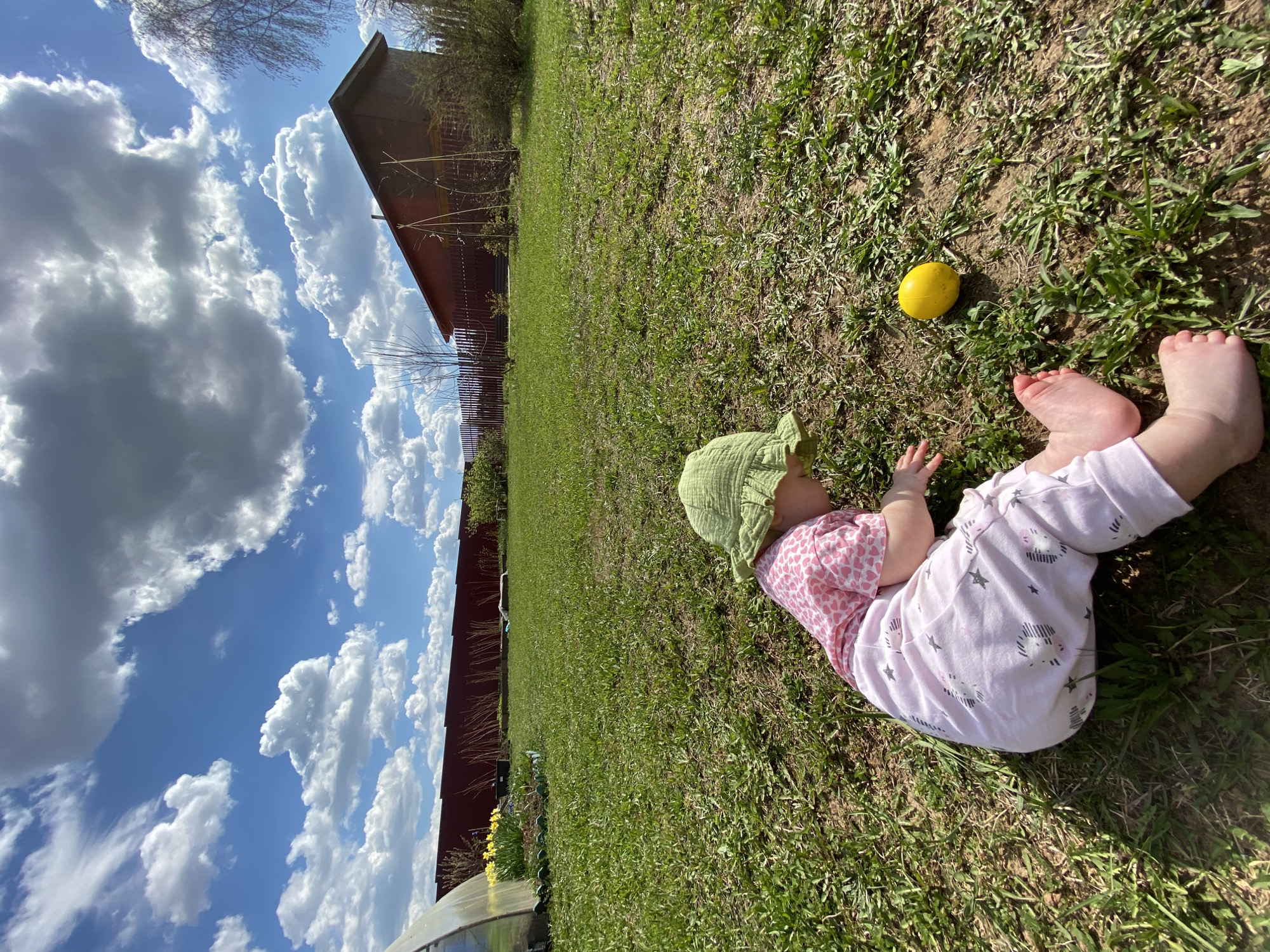 A baby wearing a green hat and pink patterned clothing lies on a grassy field next to a yellow object. The sky is visible with cumulus clouds. A red building and fence border the field. The location is Фоки, Russia.