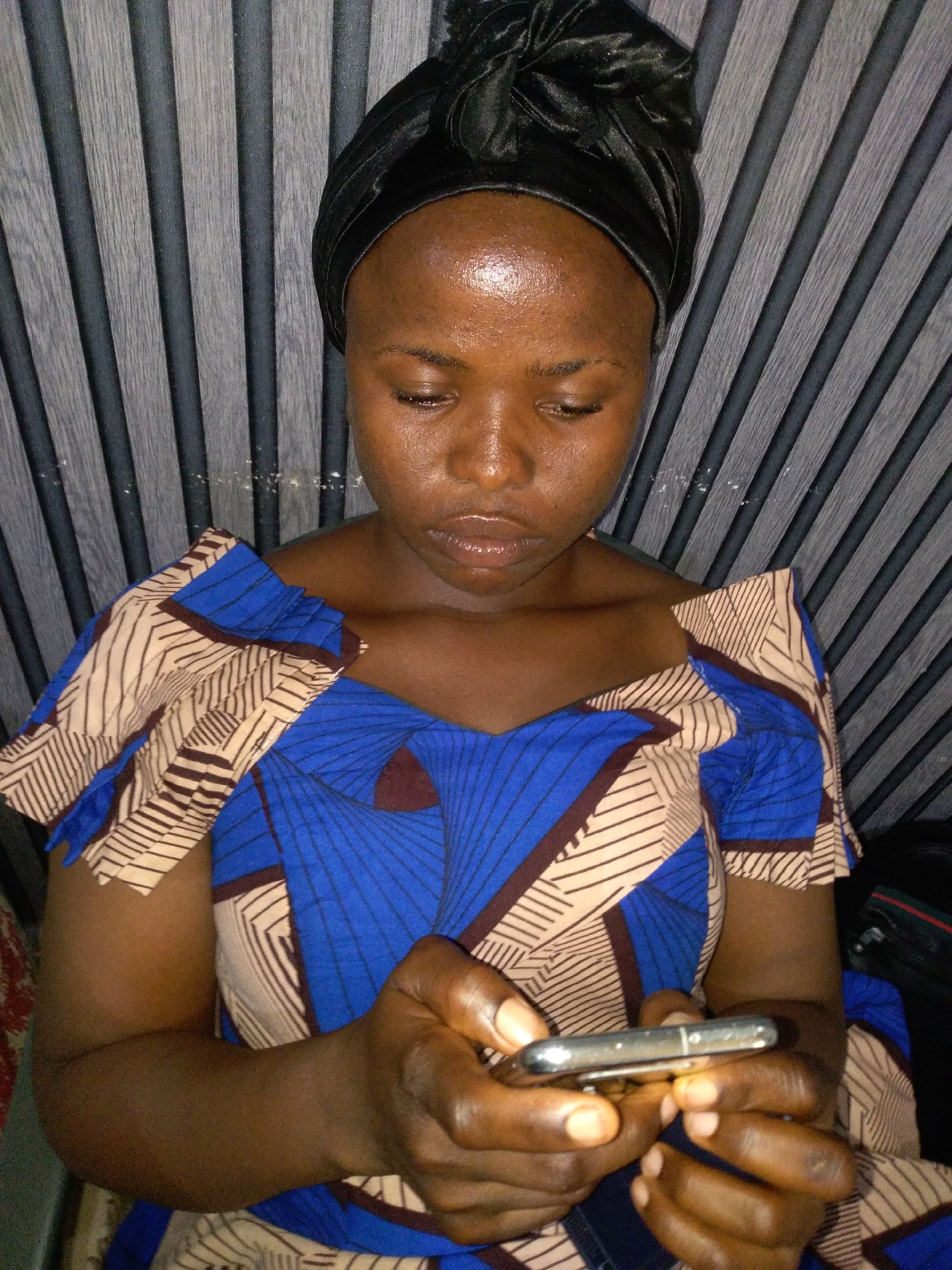 An adult female with a dark complexion is seated indoors in Unknown, Unknown, captured from the chest up. She wears a black, possibly satin, headwrap covering her hair. Her eyes are cast downwards, fixated on a silver-colored smartphone held horizontally in both hands. Her lips are full, slightly parted, and appear glossy, while her forehead exhibits a slight sheen. She is wearing a garment, likely a dress or top, patterned with large sections of royal blue featuring geometric grid designs, interspersed with tan and dark brown striped segments. The background consists of vertical wall paneling, characterized by alternating dark and lighter gray wood-grain textures, with a horizontal demarcation line visible across the upper portion.