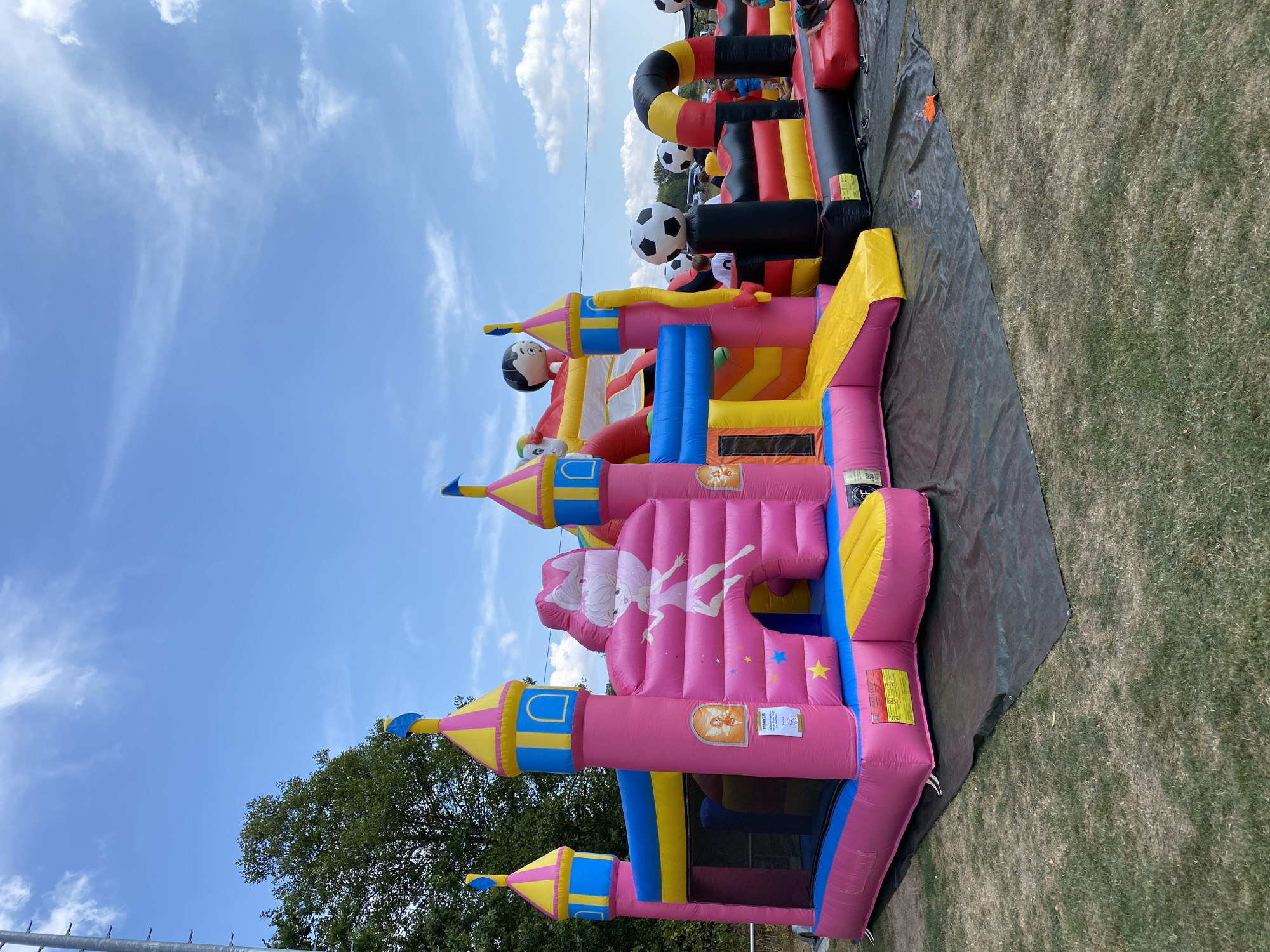 The image depicts a scene in Ingelbach, Germany, featuring multiple inflatable attractions set up on a grassy area. In the foreground, a large pink inflatable castle with turrets, windows, and a cartoon fairy character dominates the left side of the frame. It has a yellow slide and blue netting. To its right, another inflatable structure with a yellow slide and a clown face is visible. Further to the right, a black, red, and yellow striped inflatable obstacle course with soccer balls integrated into its design is present. Several children are playing or standing around these inflatables. The ground is covered with a black tarp beneath the inflatables, and dry grass surrounds the setup. The sky above is blue with scattered white clouds. A large tree and some utility poles are visible in the background.