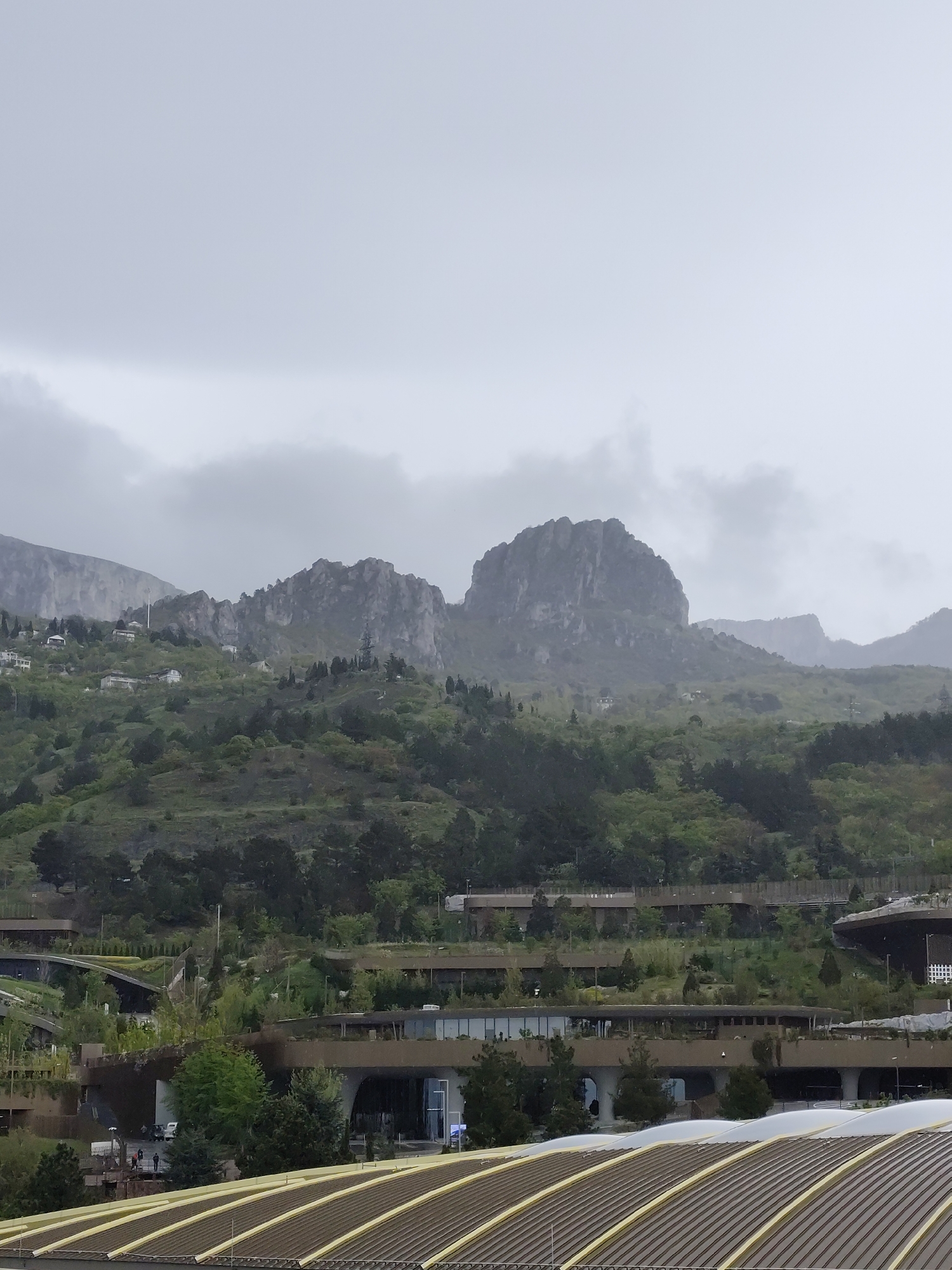 The image shows a landscape with mountains and buildings, likely in the city of Симеизский поселковый совет, Ukraine. The sky is overcast and grey. In the background are several mountains, with rocky peaks partially obscured by clouds and fog. The slopes of the mountains are covered with vegetation, including trees and shrubs, with a few scattered buildings. In the foreground, there are several low-rise buildings with flat roofs covered with greenery. The rooftops have a repeating pattern of metal beams or panels.