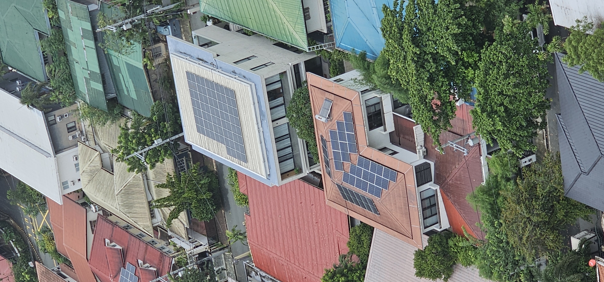 This is an aerial view of a residential neighborhood in Makati, Philippines. The image shows a cluster of houses with various roofing materials, including red tiles, green metal, and blue metal. Several houses are equipped with solar panels. Green trees and foliage are abundant throughout the area, indicating a well-maintained urban landscape. The scene depicts a typical residential street with houses, roads, and power lines.
