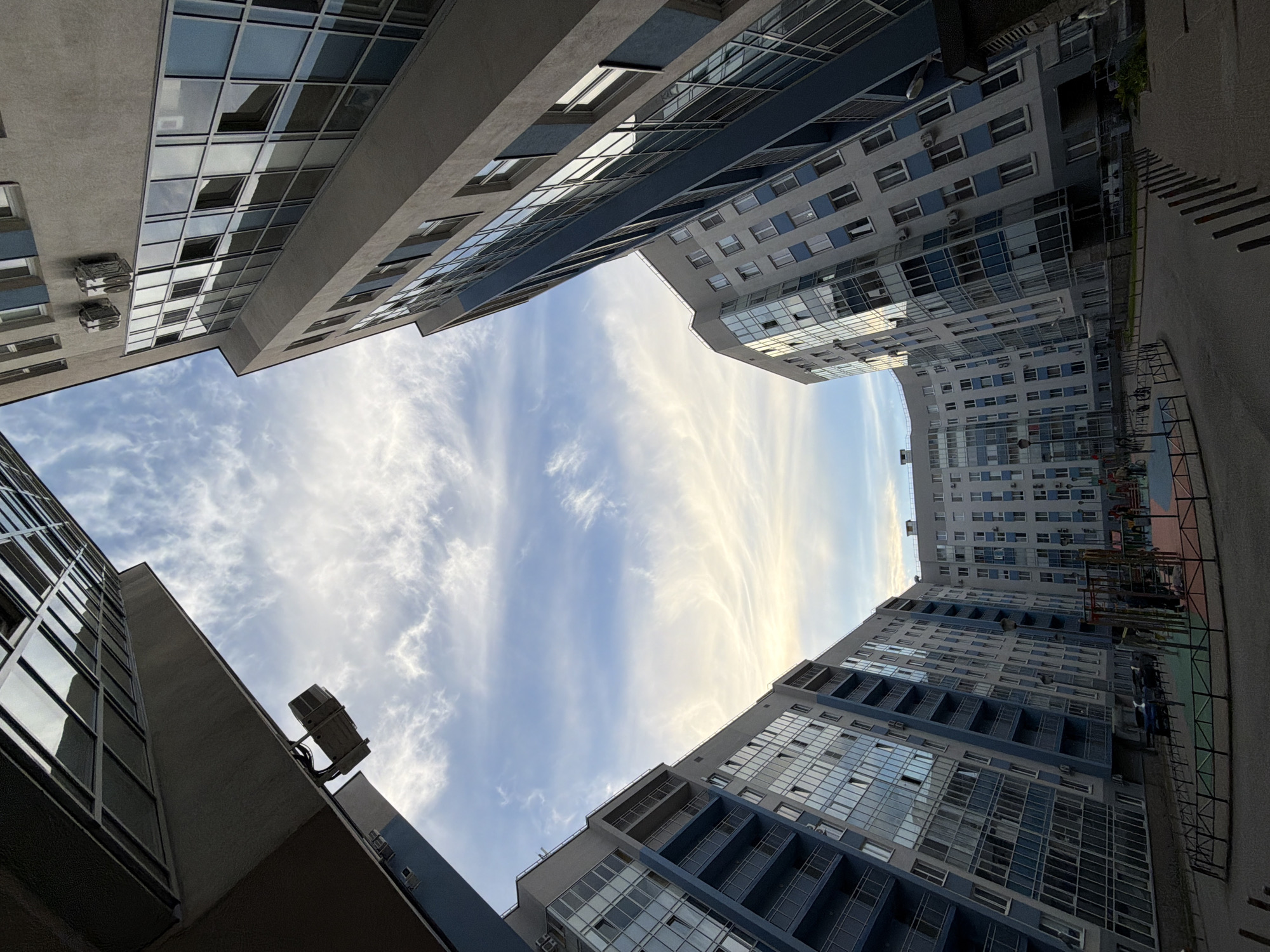 The image captures an upward-looking perspective from within a courtyard or open space surrounded by modern high-rise residential buildings. The buildings, predominantly gray and white with accents of blue/teal on balconies and facades, feature numerous windows and reflective glass panels that catch the light. Air conditioning units are visible mounted on the exterior walls of several structures. The architectural style suggests contemporary design.
The buildings frame a large expanse of sky, which is bright blue with wispy, elongated white cirrus clouds stretching across it. There's a soft, warm glow on some of the clouds, suggesting either early morning or late afternoon light. The low-angle shot creates a dramatic, converging perspective where the buildings lean inward towards the sky.
No people or visible text are present in the image. Hints from the provided crops reveal elements like paved ground, railings, and a children's playground structure in the distance, indicating a residential complex setting in Saint Petersburg, Russia. The overall scene is static, focusing on the urban architecture and the sky above.