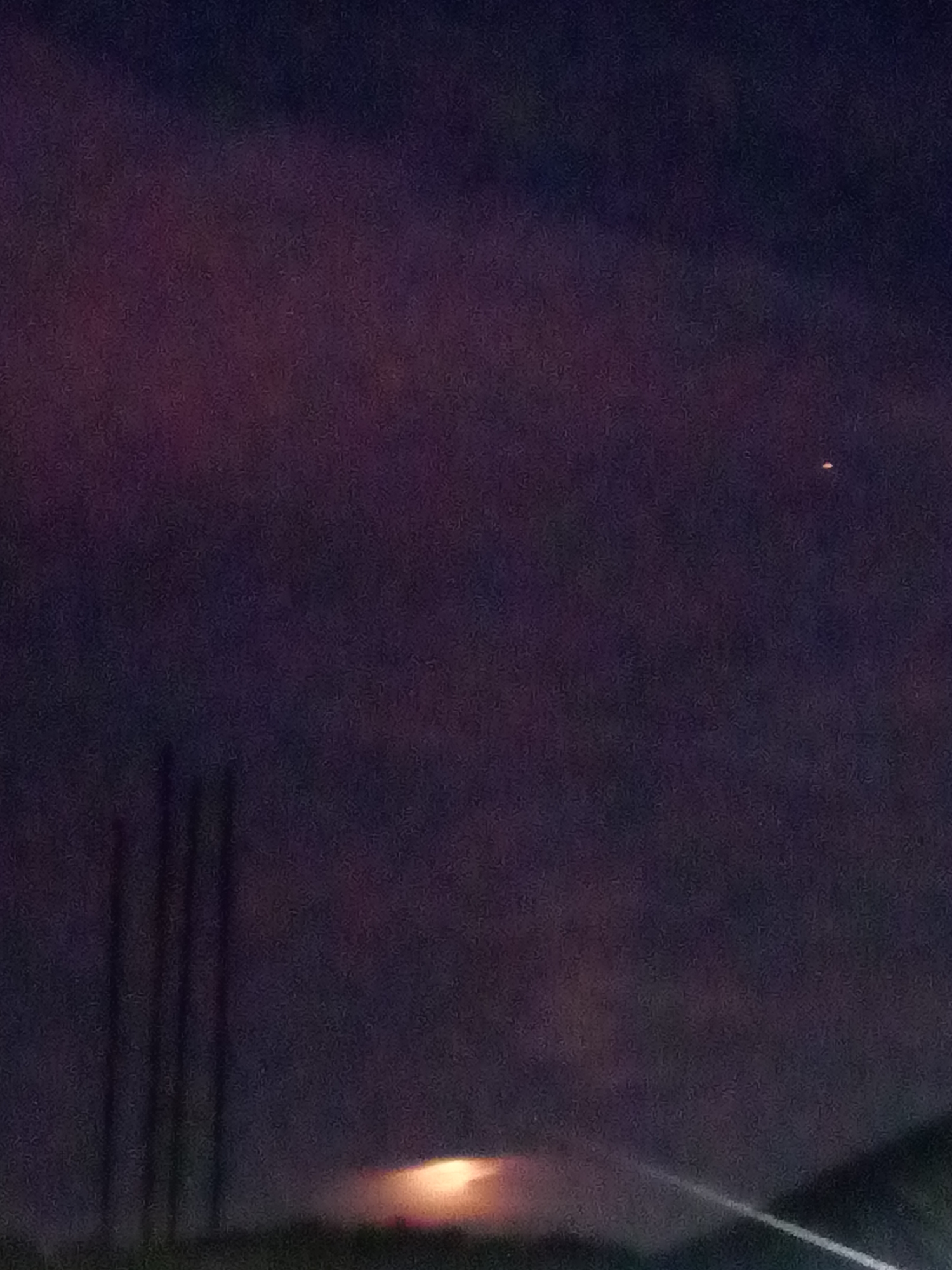 The image appears to be taken at night, possibly in or near Juan Griego, Venezuela, given the reference to the city. The scene features a dark, clouded sky with a single bright spot resembling a star or planet visible in the upper right portion. A faint, diffused light source is visible near the bottom, potentially artificial lighting or the rising moon obscured by clouds. In the lower left, dark, vertical lines suggest the presence of poles or posts. A faint line angles diagonally upward from the bottom right, potentially a road or other feature. The overall image is dark and indistinct, with minimal identifiable details.