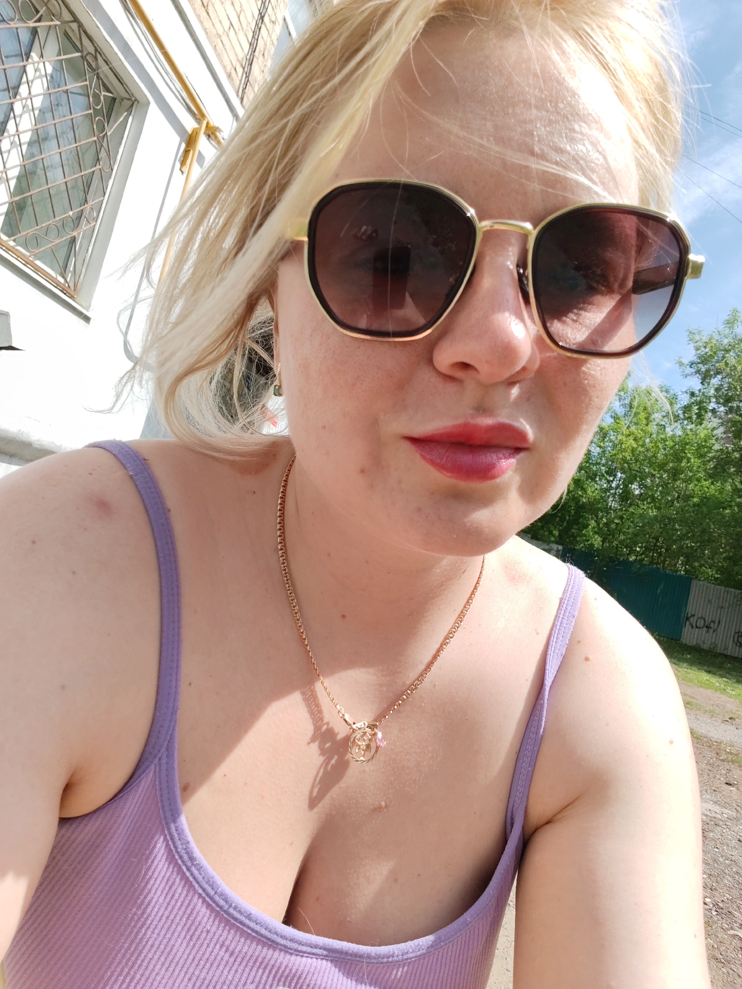 A woman with blonde hair and sunglasses takes a selfie outdoors. She is wearing a purple tank top and a gold necklace. In the background, there is a building with barred windows and some greenery. The text "K04" is faintly visible on a fence. The location is Izhevsk, Russia.