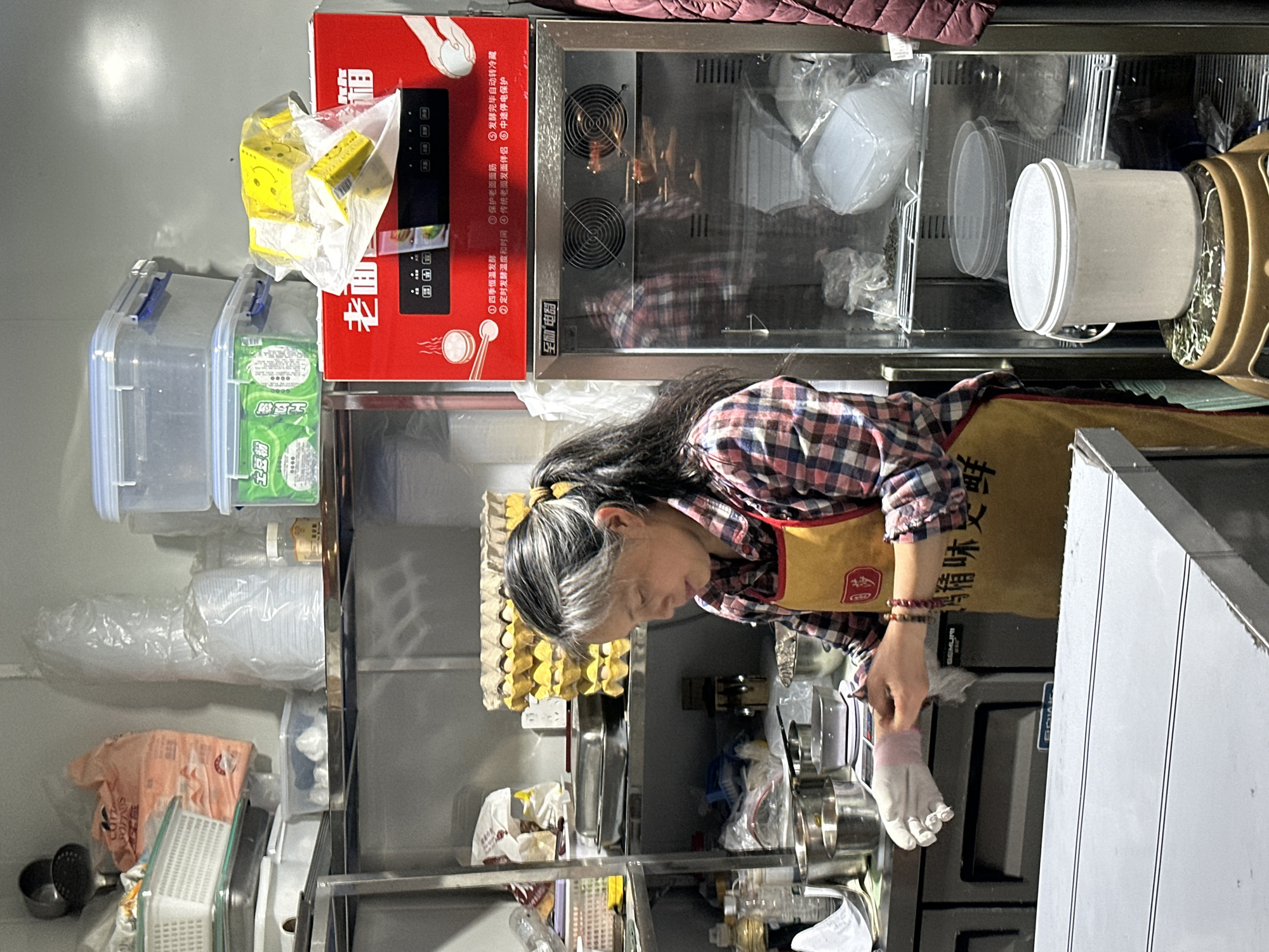 A kitchen scene shows a person wearing a plaid shirt and an apron labeled with Chinese characters and is donning a white glove to prepare food in a commercial kitchen. An assortment of eggs sits below a variety of containers and food products. Above them is a warmer. The OCR reveals Chinese characters related to food preparation. While not explicitly stated, the combination of the visible elements suggests a food business location in Jiangjin, China.