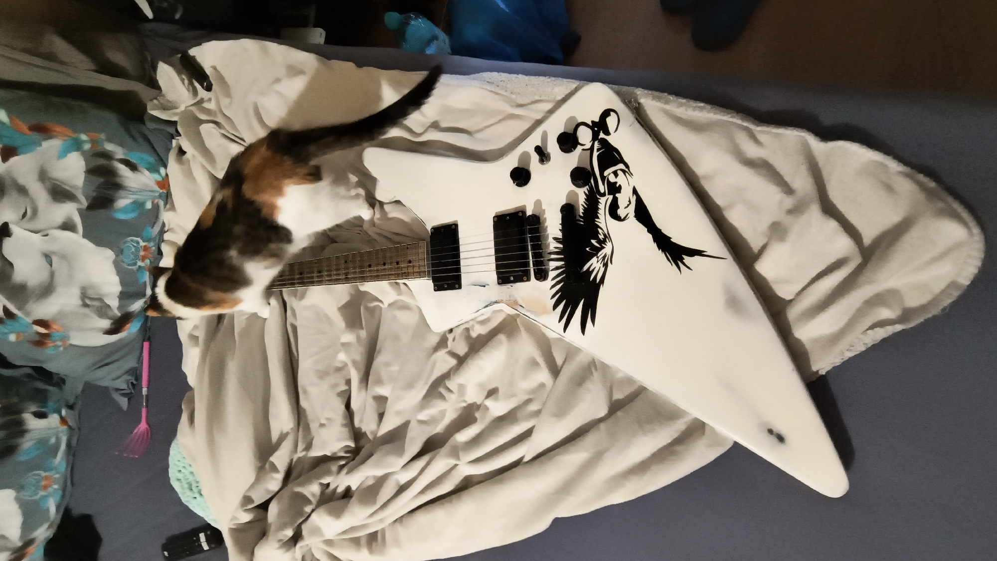 A white, electric Explorer-style guitar with a black graphic on the body lies on rumpled bedding. A calico cat stands near the neck of the guitar. The guitar is resting on top of a light-colored blanket or sheet. There is a patterned pillow, a cat toy, and a remote control nearby. The image was taken indoors. There is no evidence to suggest the location is in Sofia, Bulgaria.