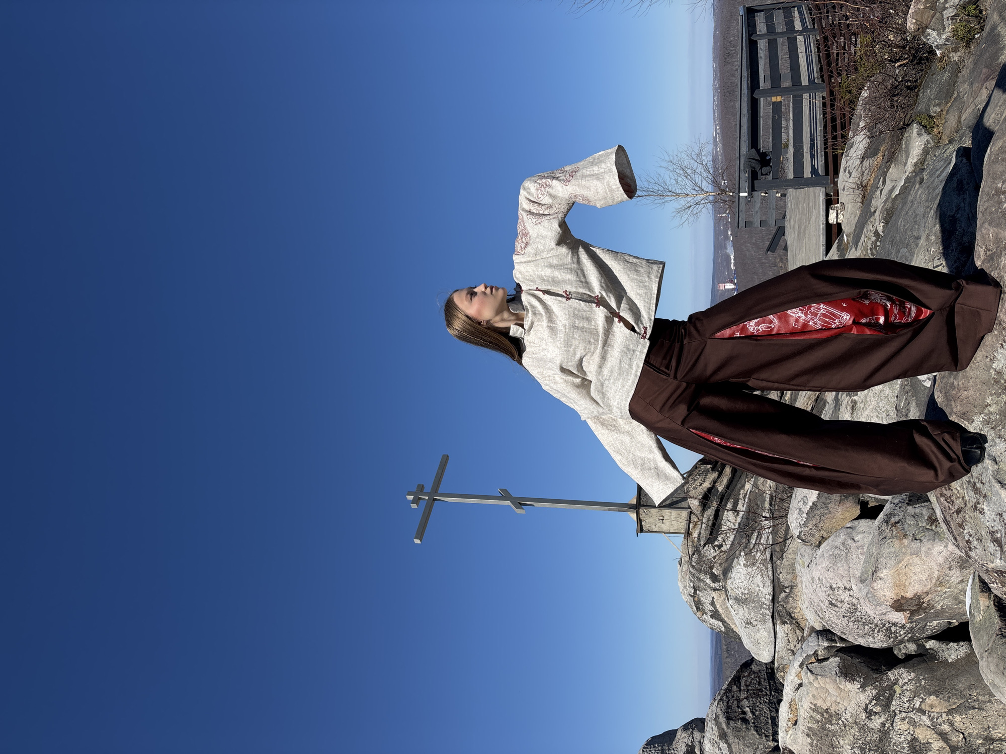 A low-angle, outdoor shot depicts a young woman standing on a rocky mountain peak in Gubakha, Russia. The sky is bright blue. She wears a light-colored, long-sleeved jacket with red trim and dark brown pants with red panels inside. Her left arm is raised, and her head is tilted back, looking upward. Behind her, there is an Orthodox cross mounted on a rocky surface. In the background, there's a railing made of dark wood and a distant view of a landscape that includes trees, terrain, and parts of what appear to be a town or city. The rock formations under the woman appear to be composed of light-colored, weathered stone.