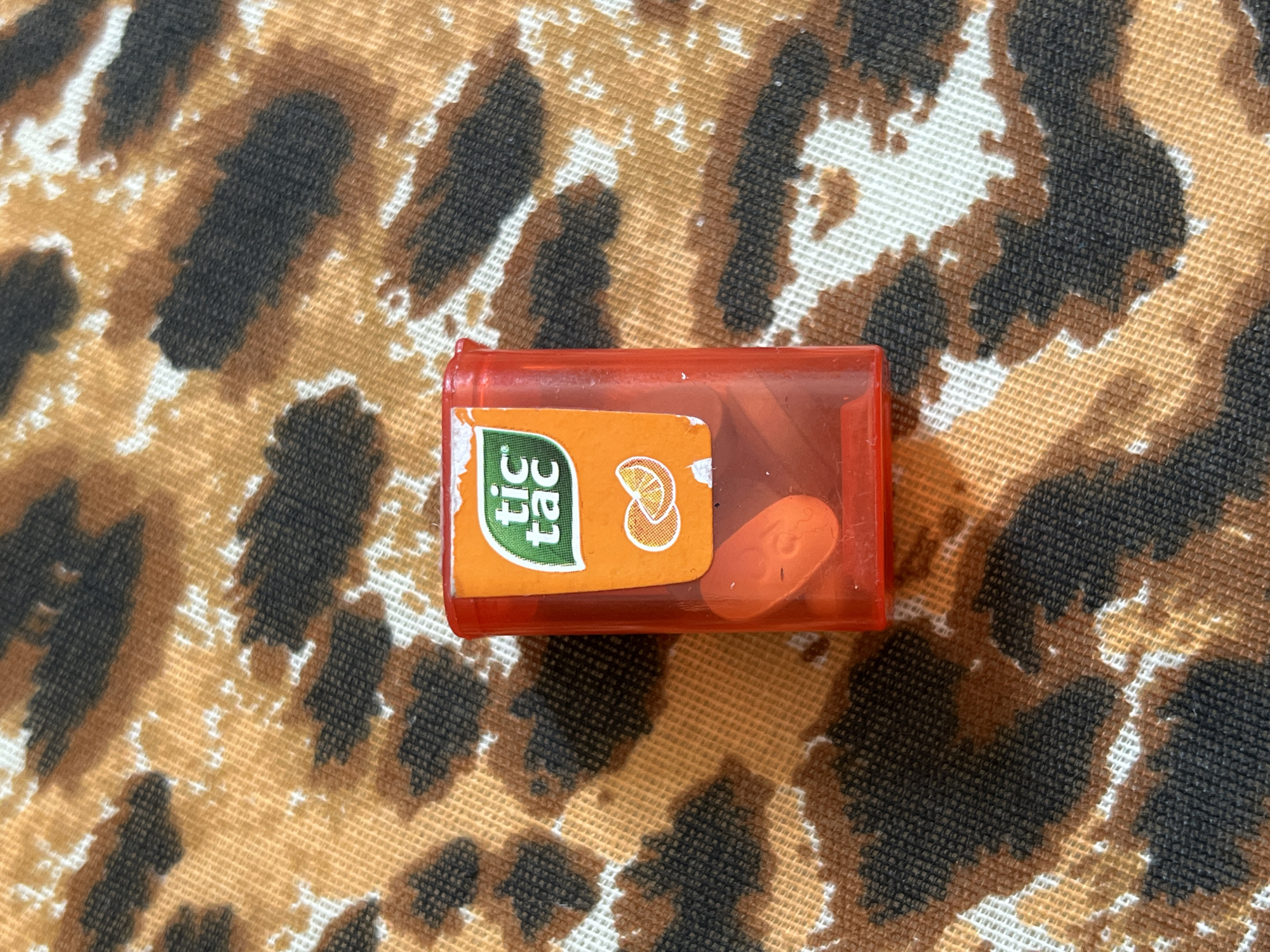 An orange Tic Tac container is centered on a surface with a leopard-print pattern. The container is partially filled with orange Tic Tacs. The photo was taken indoors. The location is not determinable.