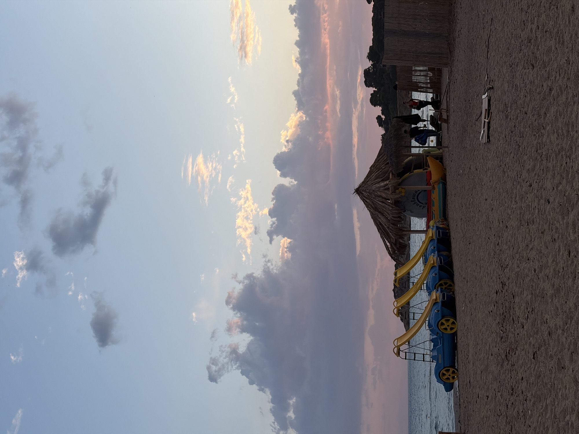 The image depicts a serene beach scene at either sunrise or sunset, characterized by a beautifully colored sky. The sky dominates the upper portion of the frame, transitioning from a light blue to soft pinks, oranges, and purples as the sun is low on the horizon. Wispy, scattered clouds in shades of grey, white, and golden-orange are dispersed across the expanse.
Below the sky, a calm body of water, likely the sea, stretches across the middle ground, reflecting the soft hues. The foreground and right side of the image feature a sandy beach with coarse sand.
Along the water's edge, several brightly colored paddle boats, designed like blue cars with yellow wheels and yellow slides, are lined up on the sand. Further back on the beach, there's a large, traditional thatched umbrella or structure, and beyond that, some wooden shacks or buildings. Near the umbrella, colorful floating devices or kayaks are visible, with one displaying the white text "double fun".
In the distance, a dark line of trees or vegetation marks the coastline. A few indistinct figures of people are faintly visible near the wooden structures and water in the background. A long, slender wooden object rests on the sand in the mid-ground. The overall atmosphere is peaceful and calm, suggesting the tranquil beauty of a seaside location in Voula, Greece, likely as the day ends.
