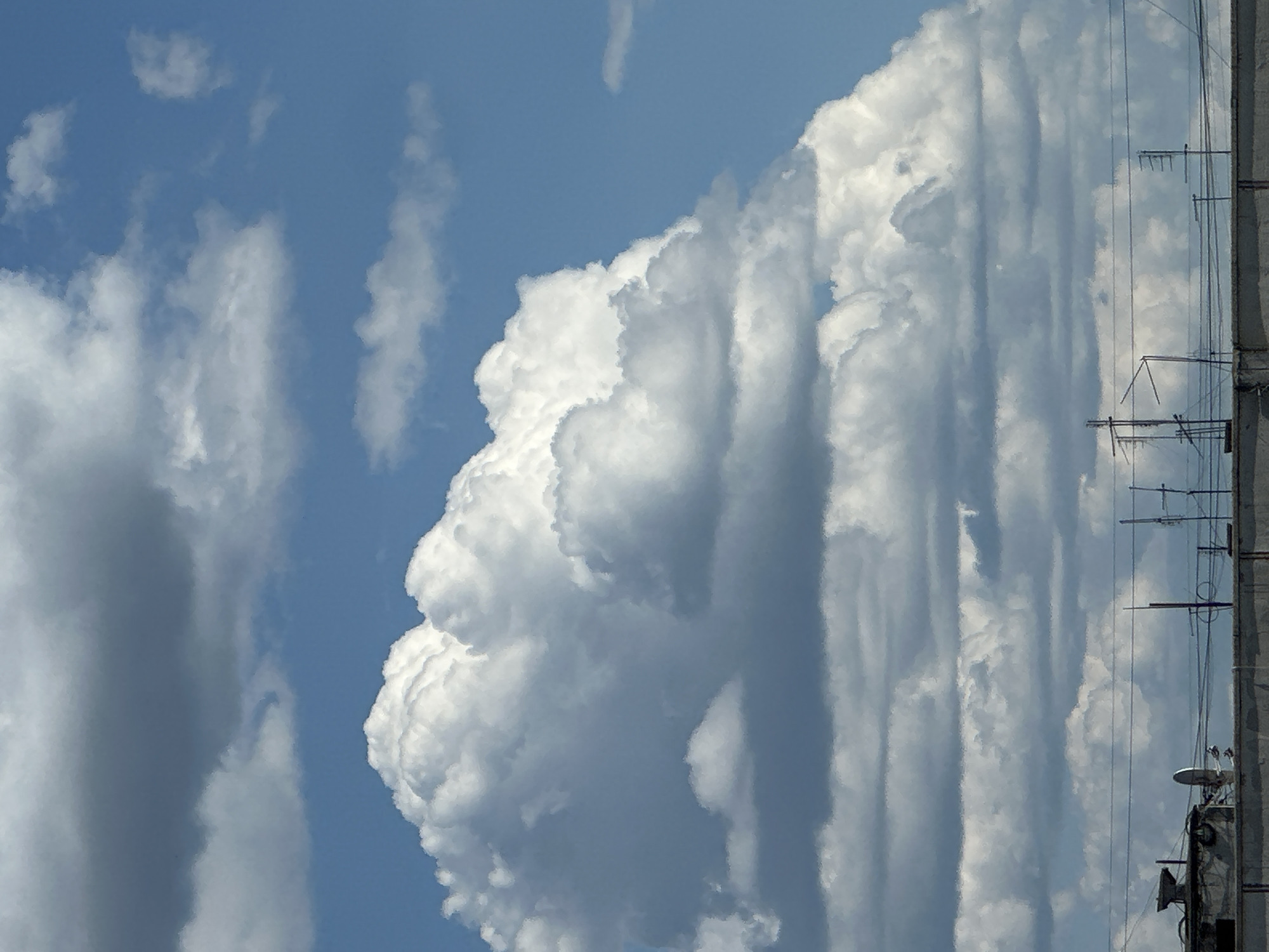 This is a vertical, eye-level medium shot of a cloudy sky with a building on the right. The sky is a light blue with various types of clouds. On the left side of the frame, there are scattered, wispy white clouds, some appearing thin and stretched. In the center and right, there is a large, dense, cumulonimbus cloud with a distinct, somewhat ominous shape. The cloud has dramatic shadowing and bright white highlights, suggesting it is illuminated by the sun. The texture of this cloud is textured and textured, with vertical striations of lighter and darker gray, giving it a powerful appearance.
On the far right of the image, a portion of a building is visible. It is a vertical structure with a concrete or stone facade. Attached to the side of the building are several antennae or telecommunication devices, appearing as a network of thin metal rods and wires. One antenna is prominent in the upper right, with multiple horizontal elements. Further down the building, smaller devices and equipment are visible, including what appears to be a satellite dish or similar object. The building occupies about one-quarter of the right side of the frame.
The lighting suggests it is daytime, likely midday or afternoon, given the brightness of the sky and the strong contrast on the clouds. The weather appears dynamic, with a large storm cloud developing or passing by, contrasted with clearer skies in other parts of the frame. There are no people or explicit signs of activity visible, but the presence of the building and antennae suggests an urban or semi-urban setting. The overall mood is one of natural power and the vastness of the sky, with a hint of technological presence from the building's equipment. There is no visible text in the image.