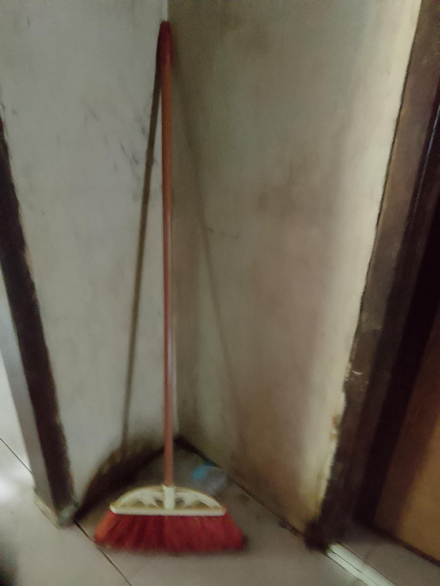 A red-bristled broom with a light-colored handle rests against a light-colored, textured wall in a corner. The broom's handle is long and slender, and it leans at an angle. The bristles are densely packed and a vibrant red. The wall appears to be a neutral tone, possibly off-white or beige, with some subtle discoloration or smudges. The scene is dimly lit, creating soft shadows. The broom is positioned in what appears to be an interior space, possibly a home or a utility area. There is no indication of the city of Merbopuntong, Indonesia, in this image.