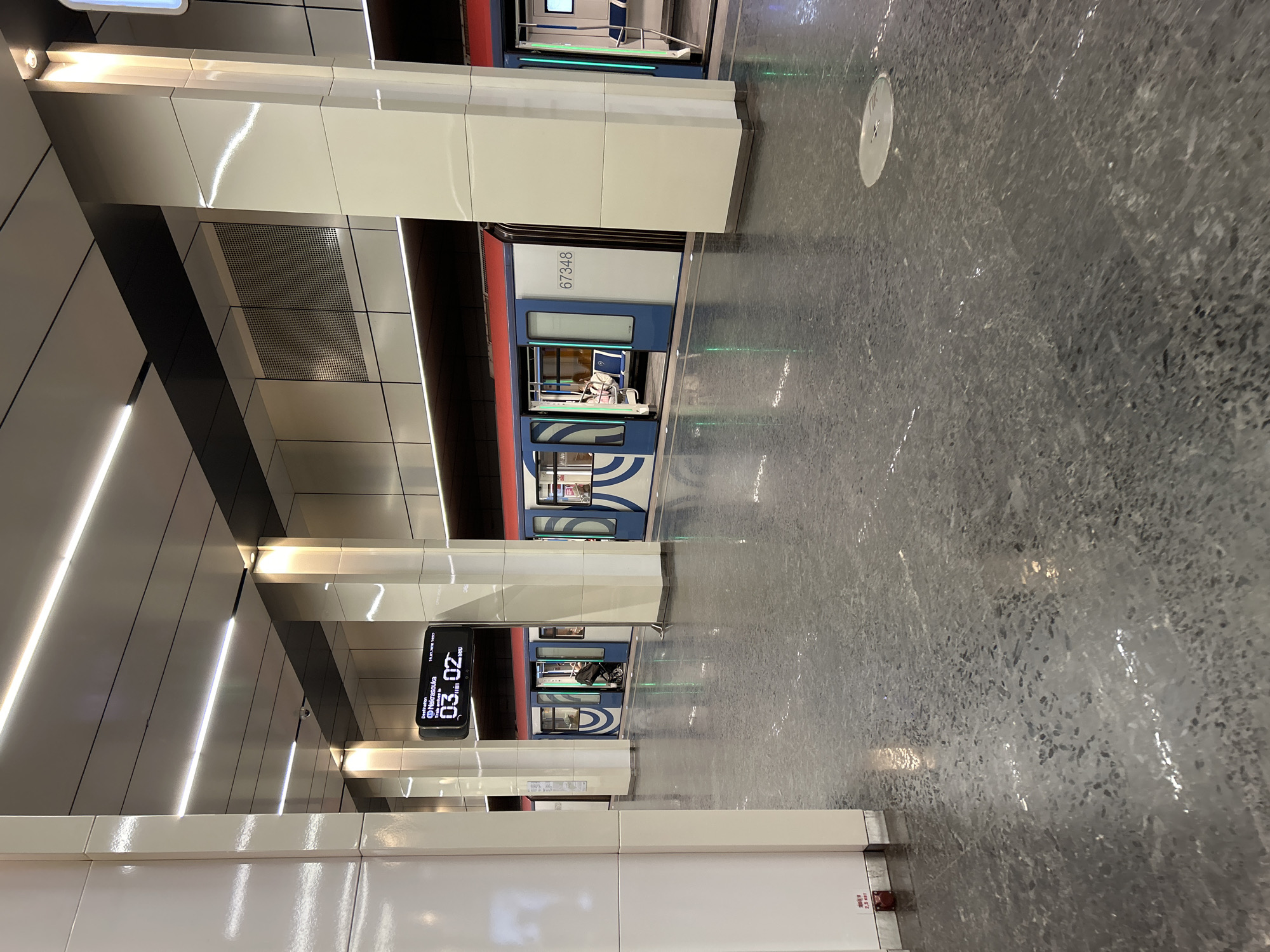 An underground subway station platform is shown with a train stopped at the platform. The station features a modern design with white tiled pillars and a grey, reflective floor. The ceiling has linear lighting. A digital display board above the train shows "Destination Nekrasovka" and the time "03.02". The train cars are blue and white with red trim, and the doors are open. Several passengers are visible inside the train, some seated, and one person with a stroller is standing near an open door. The platform is mostly empty except for the area near the train. The lighting creates reflections on the wet-looking floor.