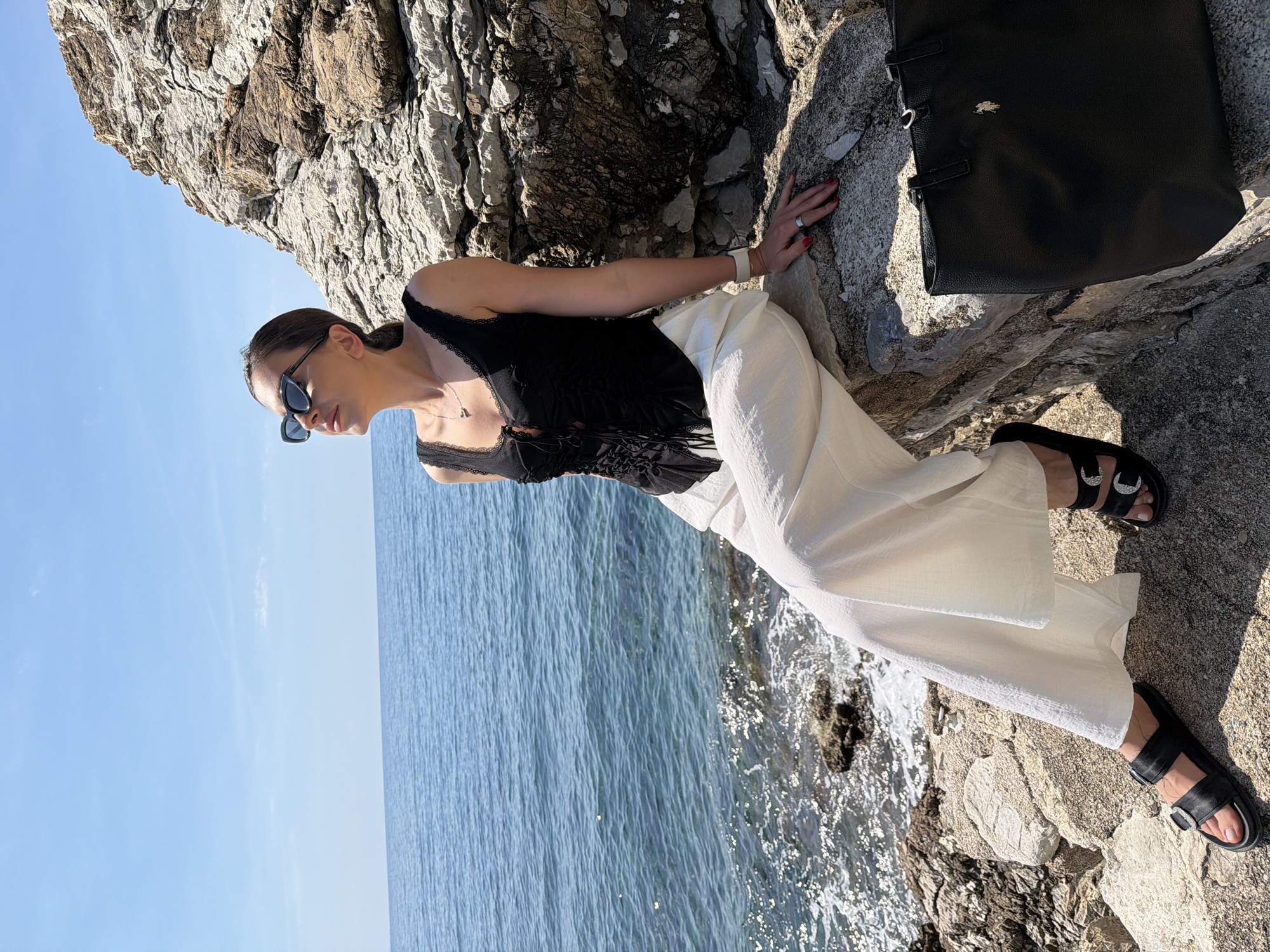 The image features a woman posing outdoors on a sunny day along a rocky coastline, specifically in Budva, Montenegro. She is leaning against light-colored, textured rocks, with her right hand resting on the stone surface.
The woman is wearing a black sleeveless top with lace trim, loose-fitting white wide-leg pants, and black open-toed sandals with distinctive silver-toned buckles. She has sunglasses on, a necklace with a small pendant, a watch on her left wrist, and a ring on her right hand, where red nail polish is visible. Her hair appears to be pulled back.
Behind and to her left, a clear blue sky meets a calm, vast expanse of blue sea with small ripples. The water gently laps against the rocky shore where she is seated. The bright sunlight casts distinct shadows, indicating a midday or early afternoon setting. To her right, a black tote bag with a small golden animal-shaped logo is placed on the rocks. The scene conveys a relaxed coastal atmosphere. No visible text is clearly discernible on clothing or accessories beyond the bag's logo.