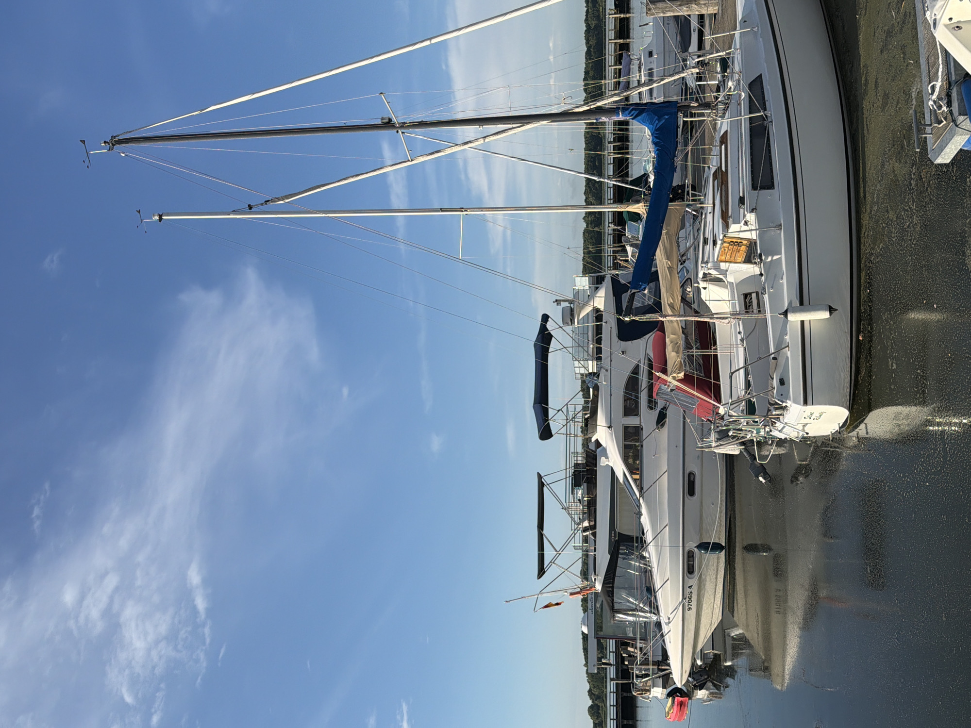 This media file captures a scene of several sailboats docked in a marina under a clear blue sky with wispy white clouds. The image is oriented vertically, with the boats stretching from the bottom of the frame upwards. The sun appears to be shining brightly, suggesting it is either morning or afternoon. The water in the marina is calm and reflective, showing the undersides of the boats.
The main subject is a fleet of sailboats, predominantly white with dark blue sails and accents. The masts of the sailboats are prominent, reaching high into the sky, with rigging and sails visible. One sailboat in the foreground has a blue sail partially unfurled and a white hull with dark trim. Another sailboat behind it also features white hulls and dark blue covers. The boats are moored next to a wooden dock.
Notable details include what appear to be wind instruments at the top of the masts. There is some greenery visible in the background, suggesting a landmass or shore beyond the marina. The overall atmosphere is one of tranquility and a beautiful day for sailing.
Visible text includes "97065-A" on the hull of one of the sailboats. Another sailboat has "Sirens Call" handwritten on its stern. The location context provided indicates this scene is in Ribnitz-Damgarten, Germany.